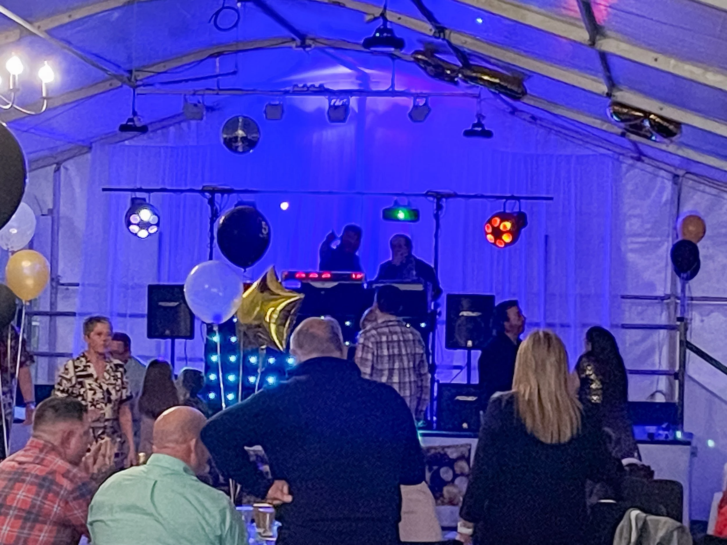 People at a party or celebration event in a tent with music and balloons, some dancing and socializing under colorful lights.