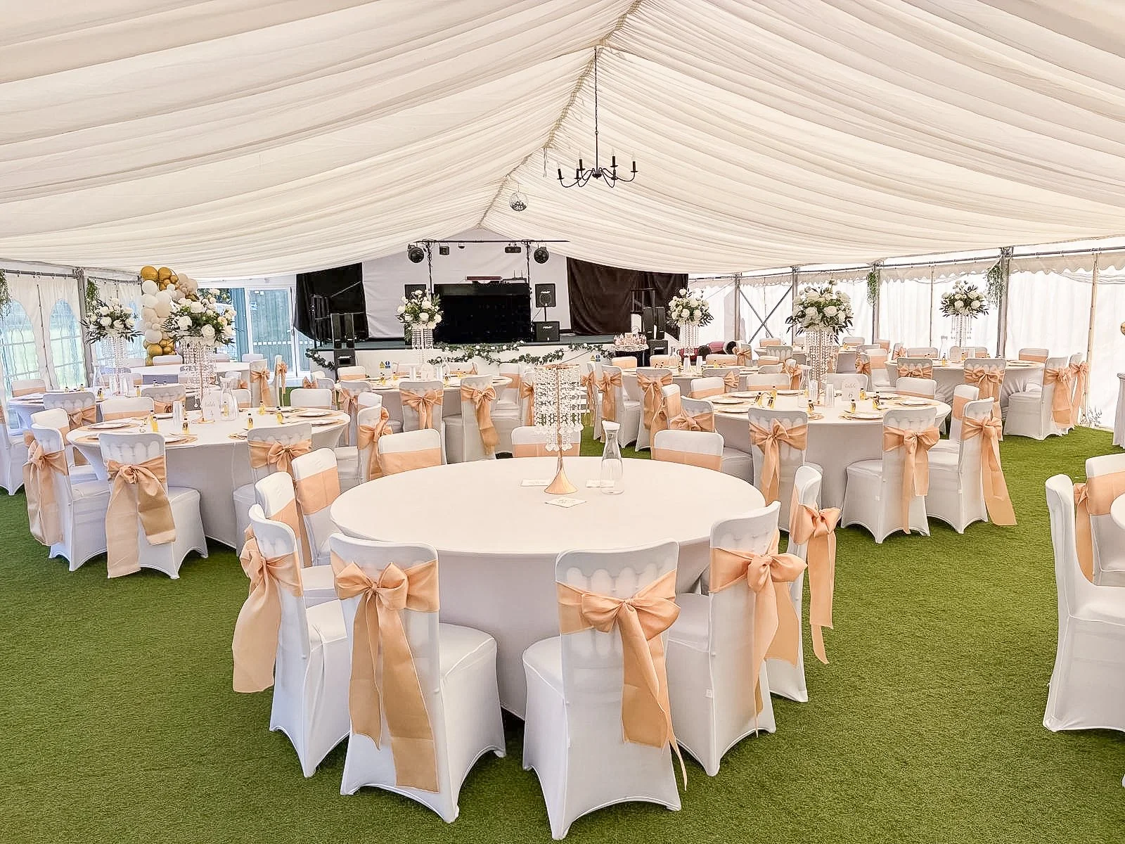 Elegant event tent with round tables adorned with white tablecloths, peach-colored bows on chairs, floral centerpieces, and a stage at the back with sound equipment and a large screen.