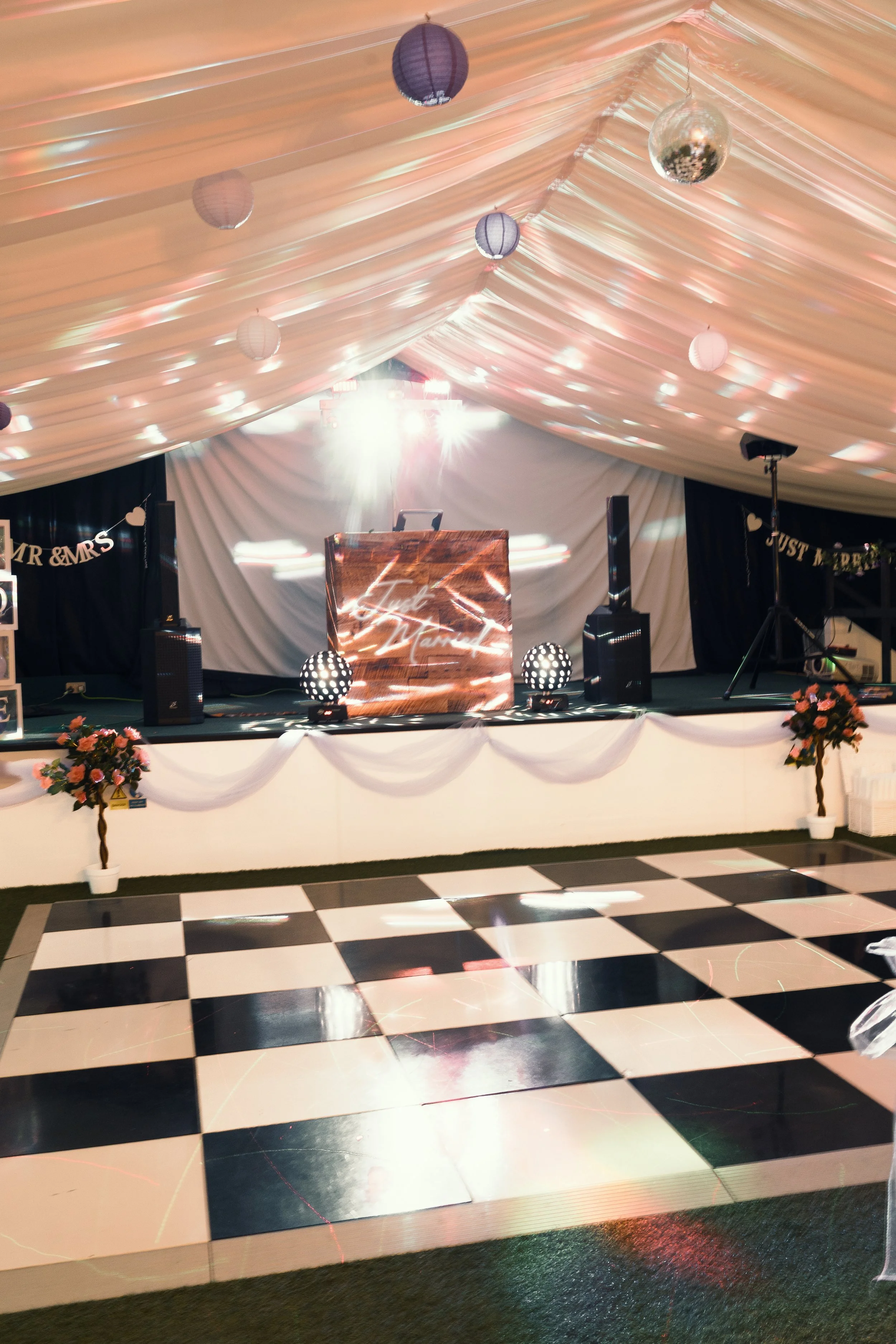 Wedding reception dance floor with a decorated stage and a checkered black and white dance floor in front. The stage has floral arrangements and a screen with the name 'Luz & Manuel,' and is set under a fabric canopy with hanging spherical lights and