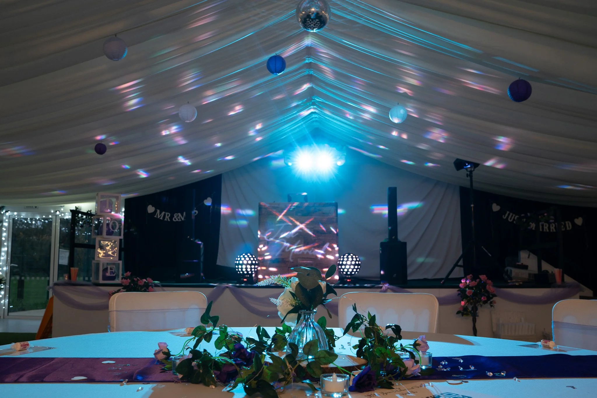 Decorated event space with a stage, LED screen, and lighting effects, set for a celebration with floral arrangements and candles on the table.