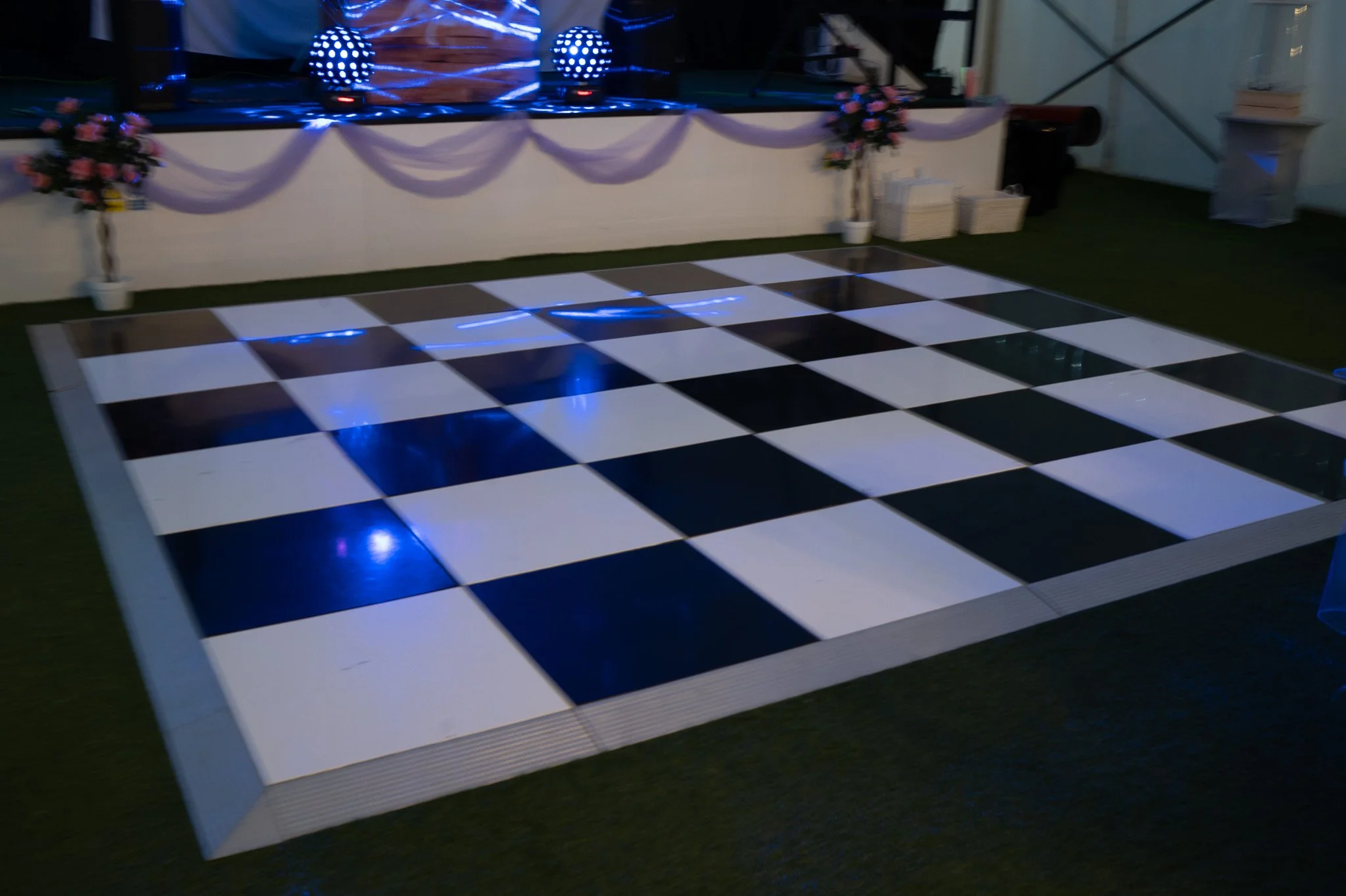 Empty black and white checkered dance floor with purple drapes, floral arrangements, and stage lighting in a decorated event space.