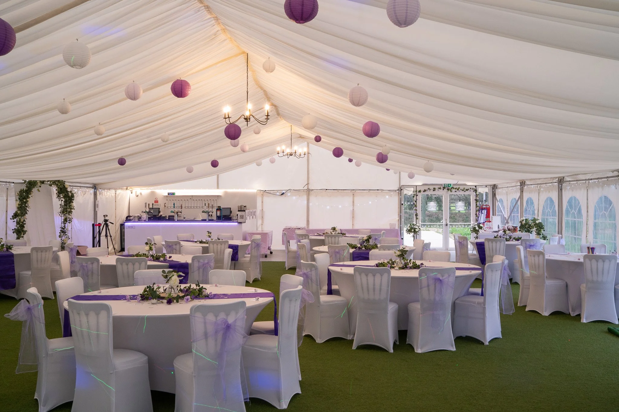 Decorated wedding reception tent with white draped ceiling, purple and white paper lanterns, round tables with white chair covers and purple sashes, floral centerpieces, a bar area, and double glass doors with greenery outside.