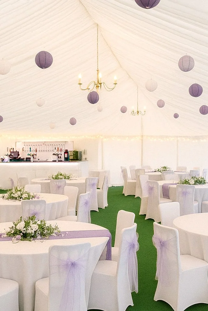 Wedding reception tent decorated with white draped fabric, purple paper lanterns, gold chandeliers, and round tables with white chairs and floral centerpieces.