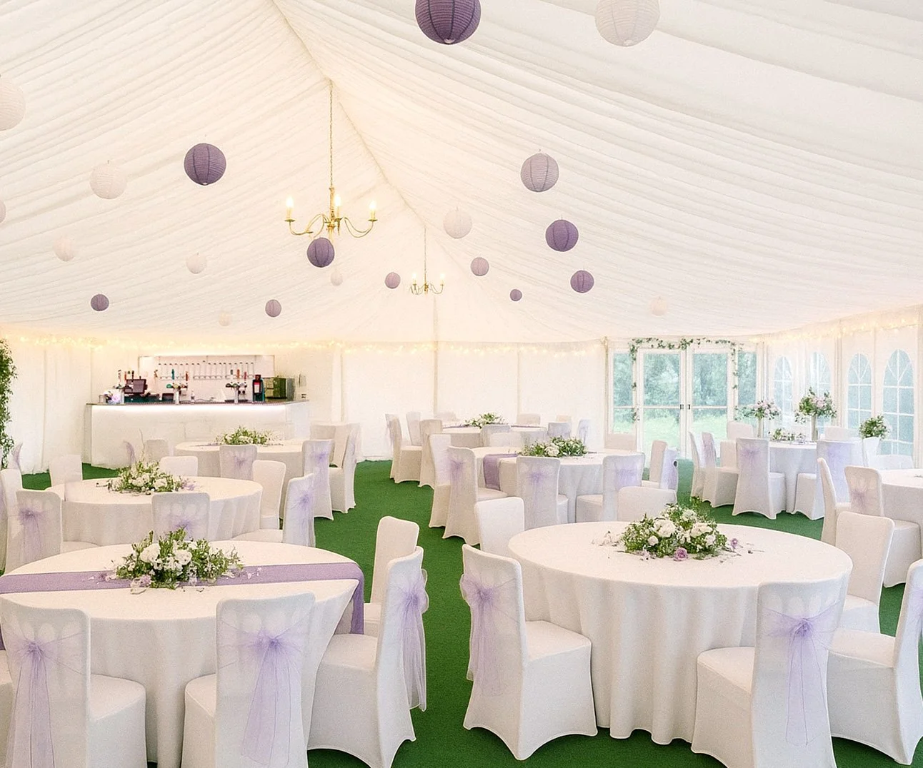 Elegant event tent decorated with purple and white paper lanterns, chandeliers, floral centerpieces, and round tables with white chairs covered in white fabric with purple bows. The interior has green carpet flooring and large windows.