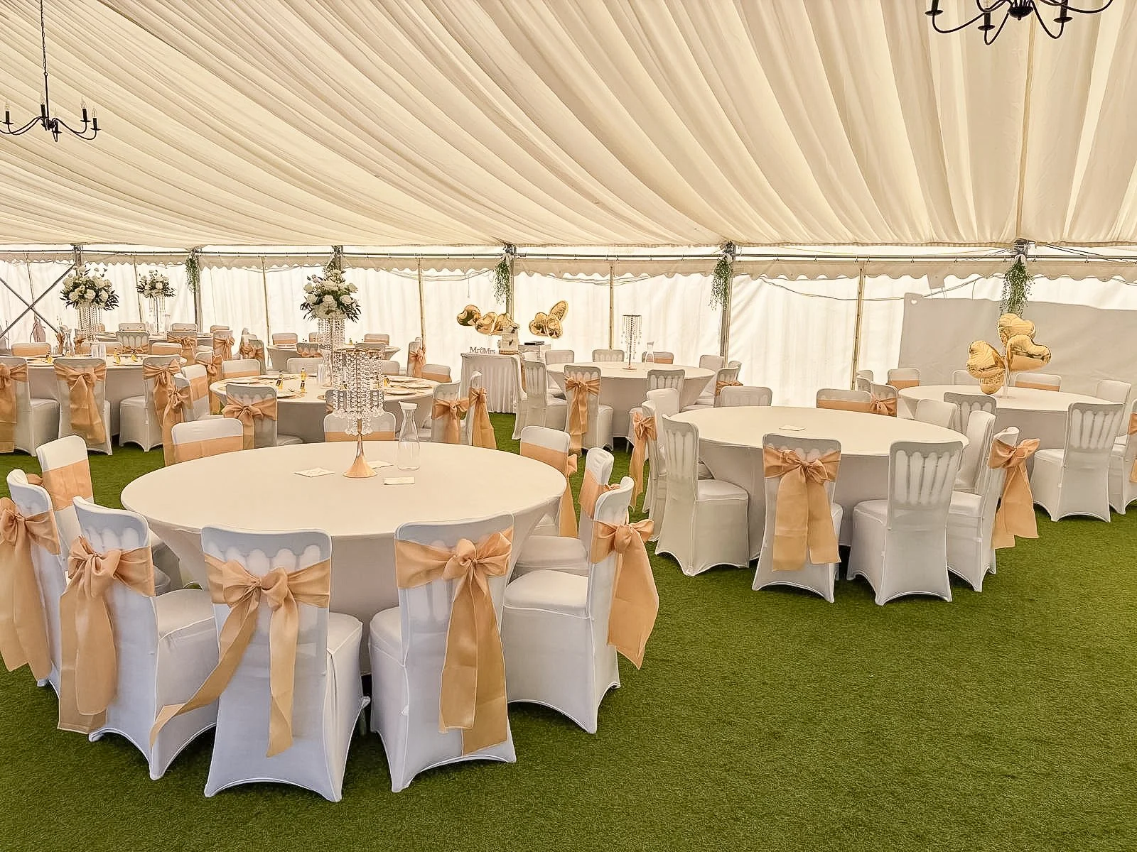 Wedding reception inside a large tent with round tables decorated with white tablecloths, peach ribbons on chairs, floral centerpieces, and balloons.