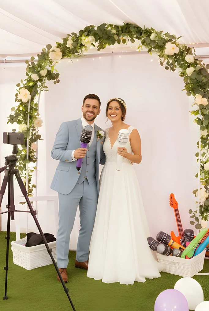Bride and groom smiling at camera in photobooth area under a floral arch, holding microphones, at a wedding reception.