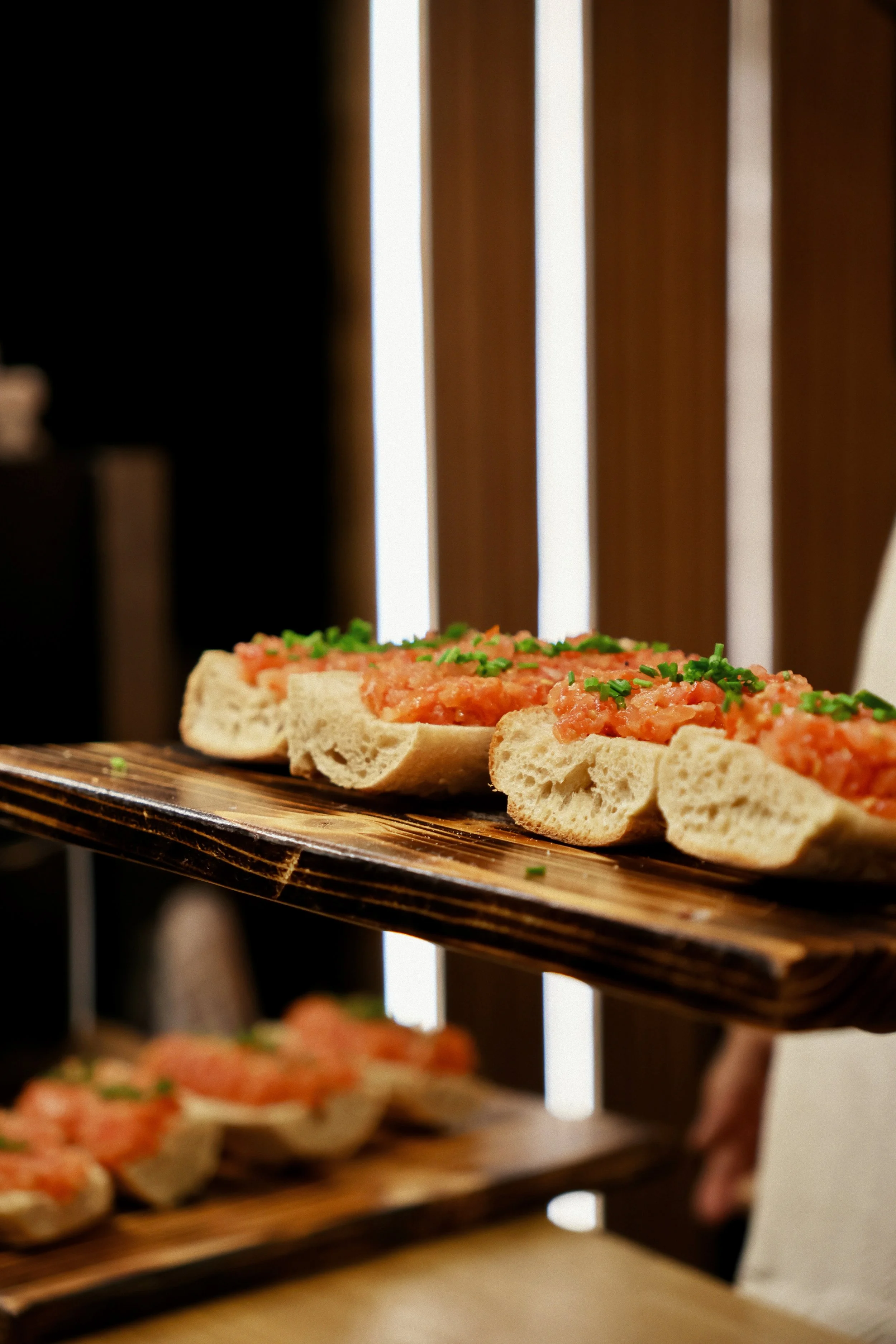 Canapés à base de pain, garnis de saumon ou de tartare de saumon, décorés de ciboulette, présentés sur une planche en bois.