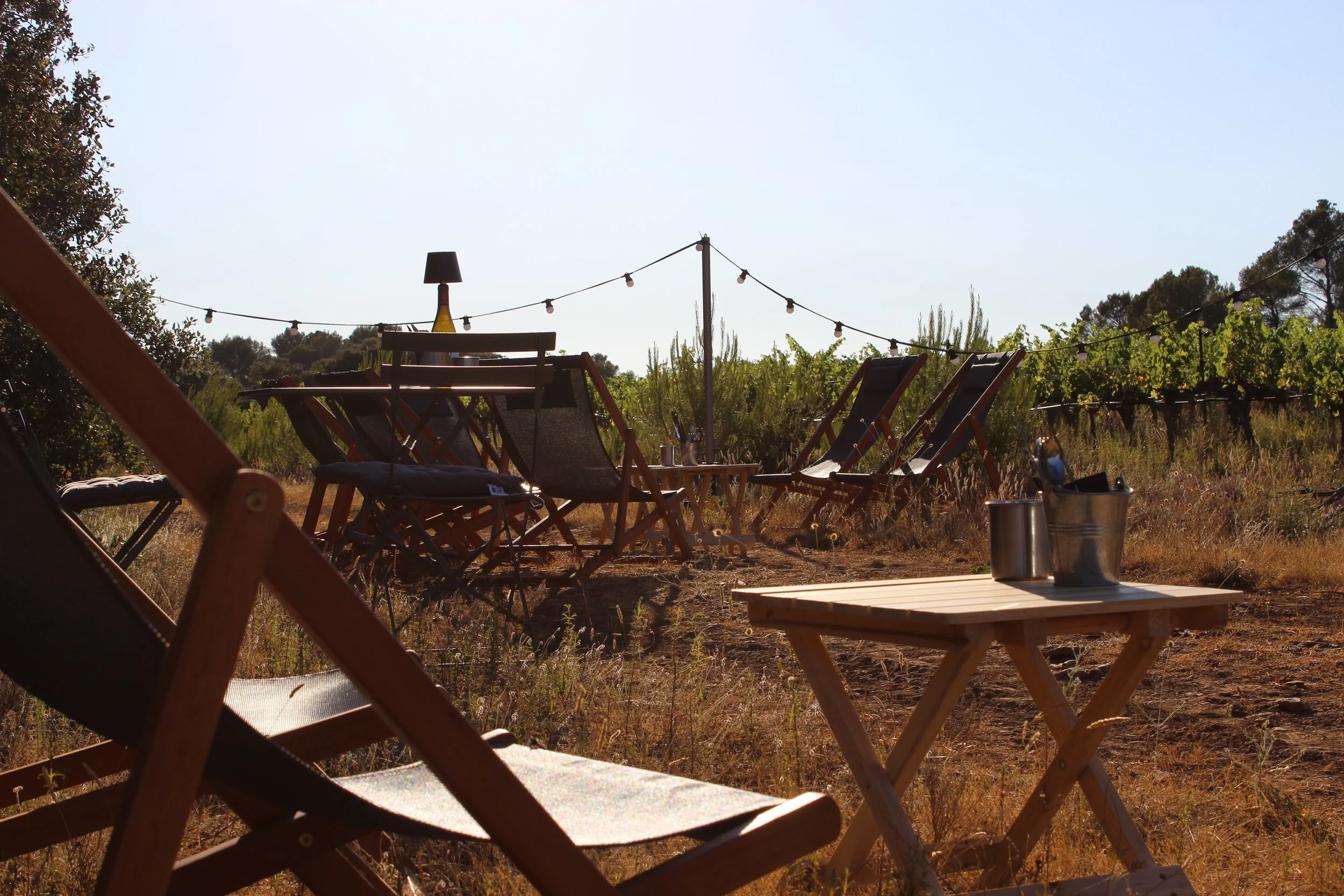 Chaises pliantes et une table en bois posée dans un champ en plein air, avec des lumières de fête suspendues et une bouteille sur la table, dans un paysage rural ensoleillé.