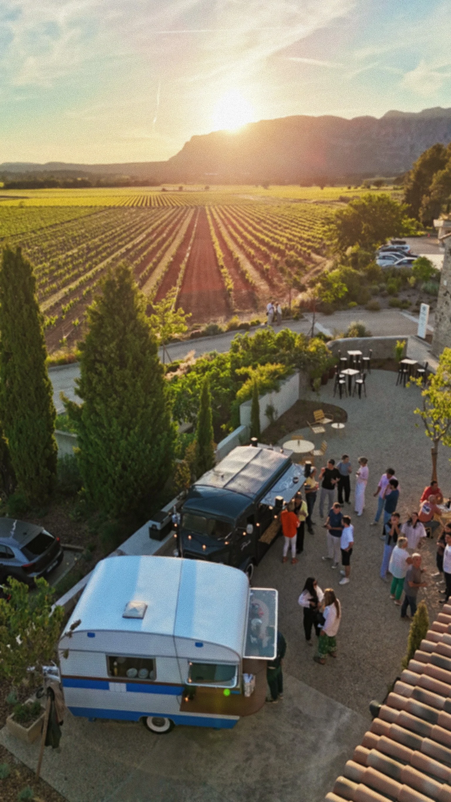 Une vue d'une fête en plein air avec un groupe de personnes près d'une caravane et d'un food truck, avec un vignoble et une montagne en arrière-plan.