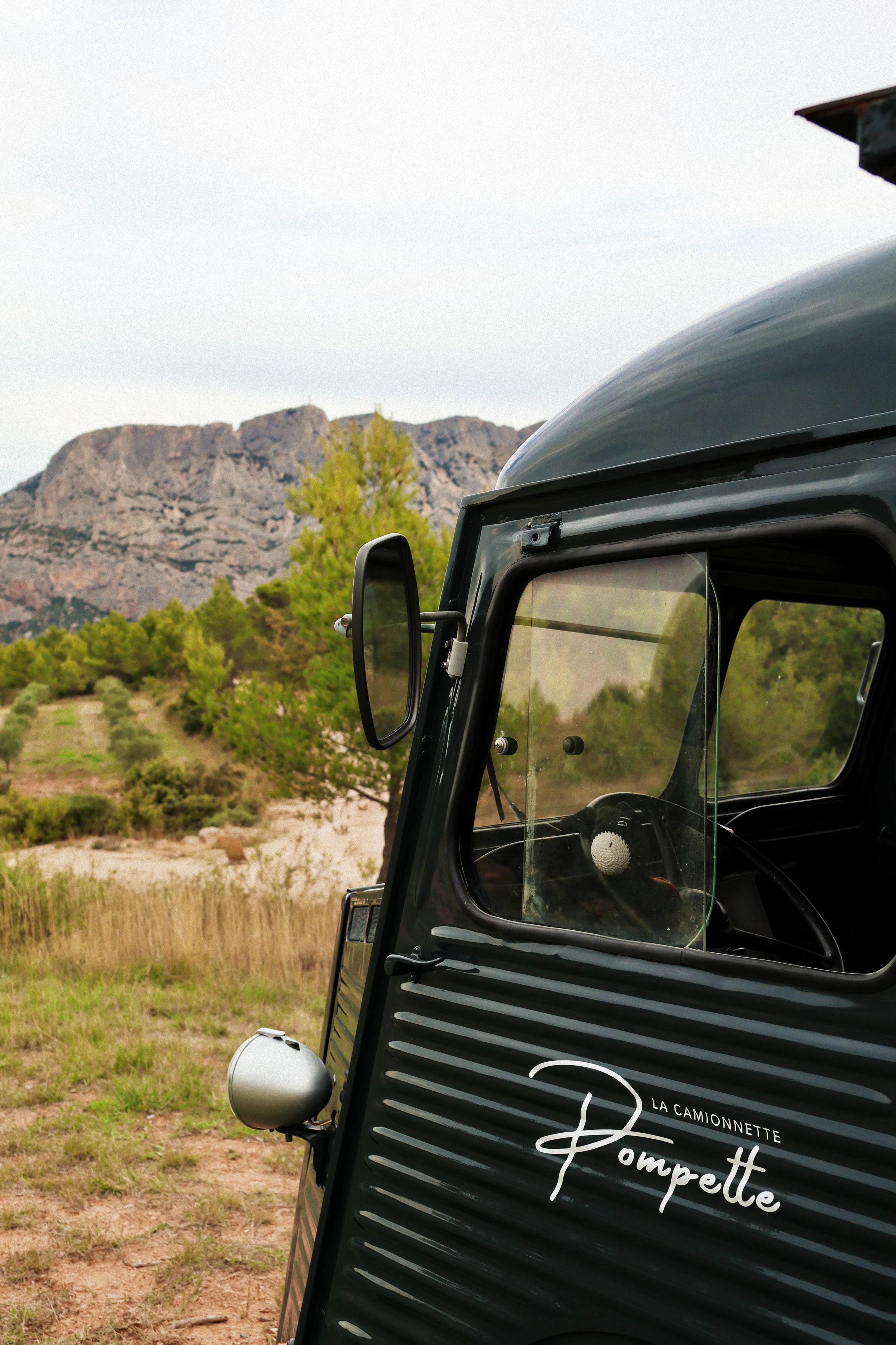 Une petite camionnette noire appelée 'La Camionnette Pompette', stationnée dans un paysage avec des arbres verts et des montagnes en arrière-plan.