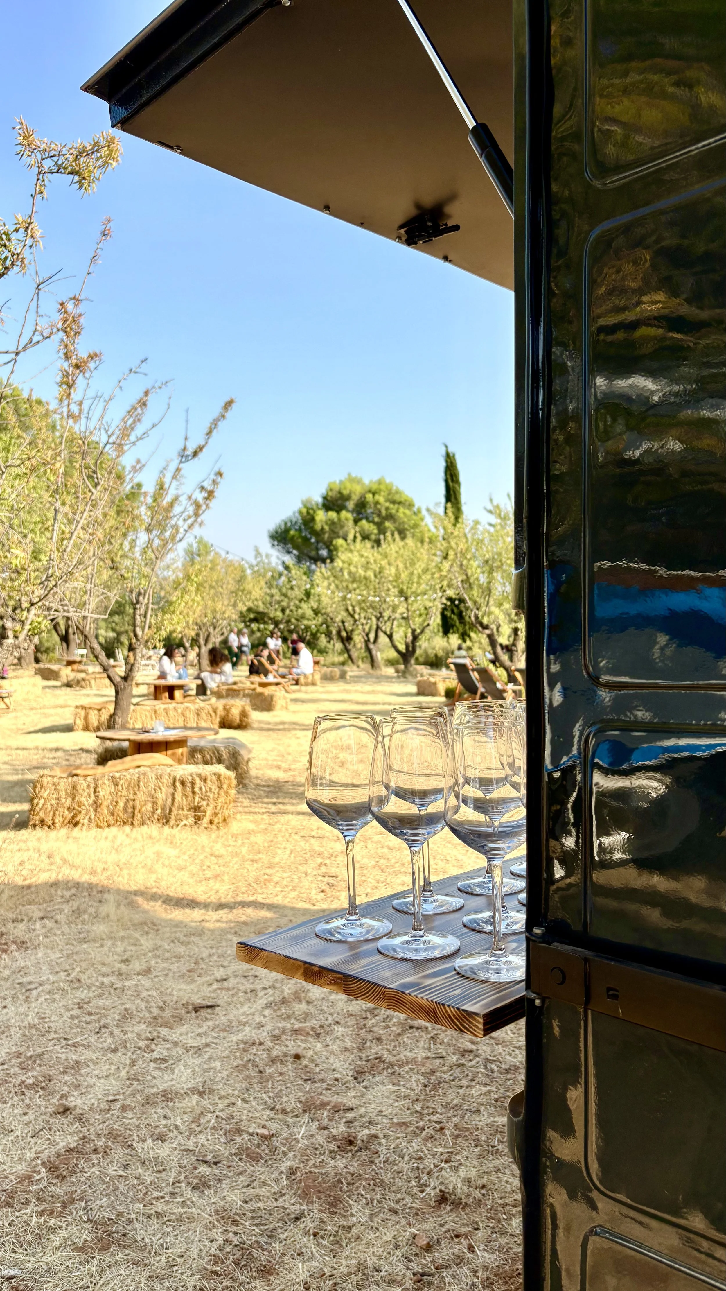 Verre à vin sur une table en bois dans une fête en plein air avec des arbres et des personnes assises en arrière-plan.