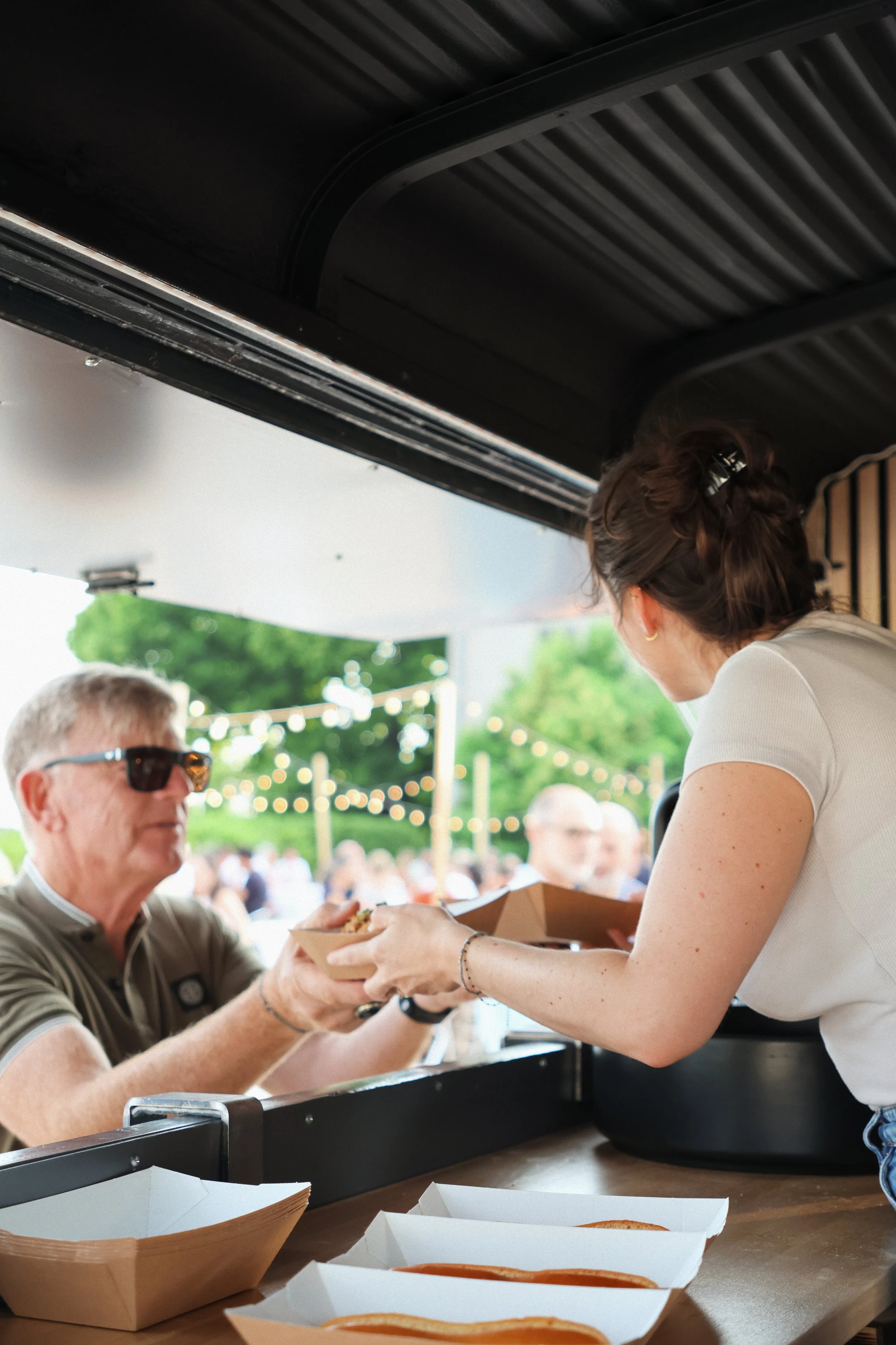 Une serveuse tend un plat à un client lors d'un événement en plein air avec des lumières suspendues et de nombreux visiteurs en arrière-plan.