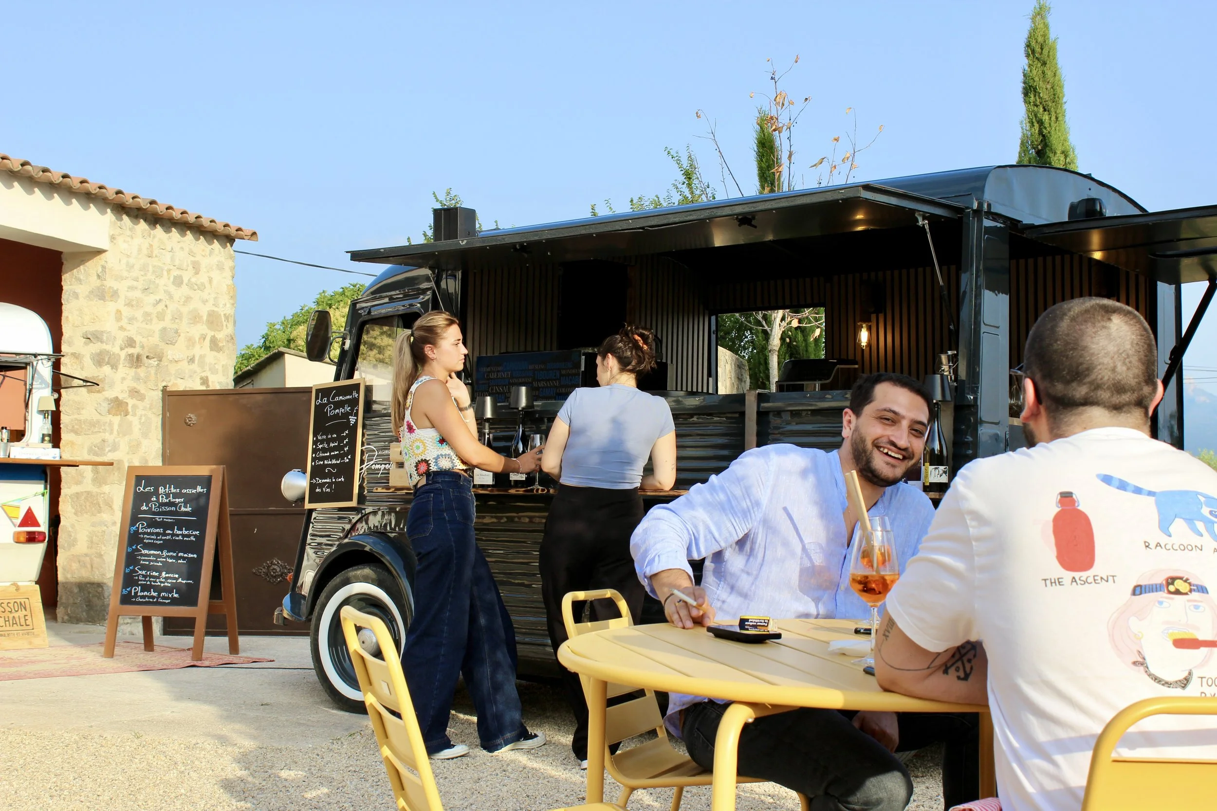 Des gens souriants qui mangent et socialisent devant un food truck en plein air, avec un menu écrit sur un tableau noir et un décor ensoleillé.