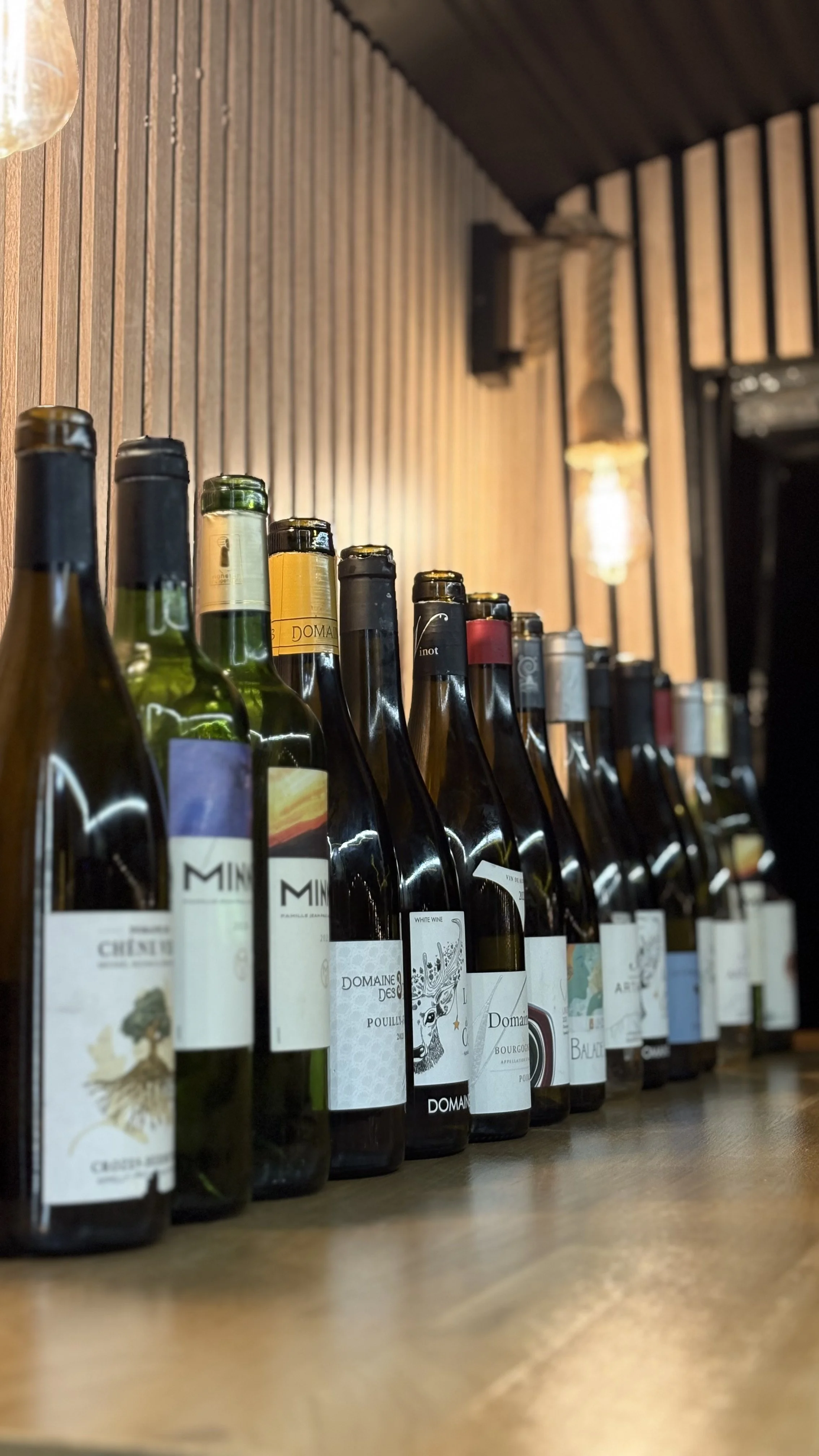 Une rangée de bouteilles de vin alignées sur une surface en bois dans un intérieur avec un mur en bois et un luminaire suspendu