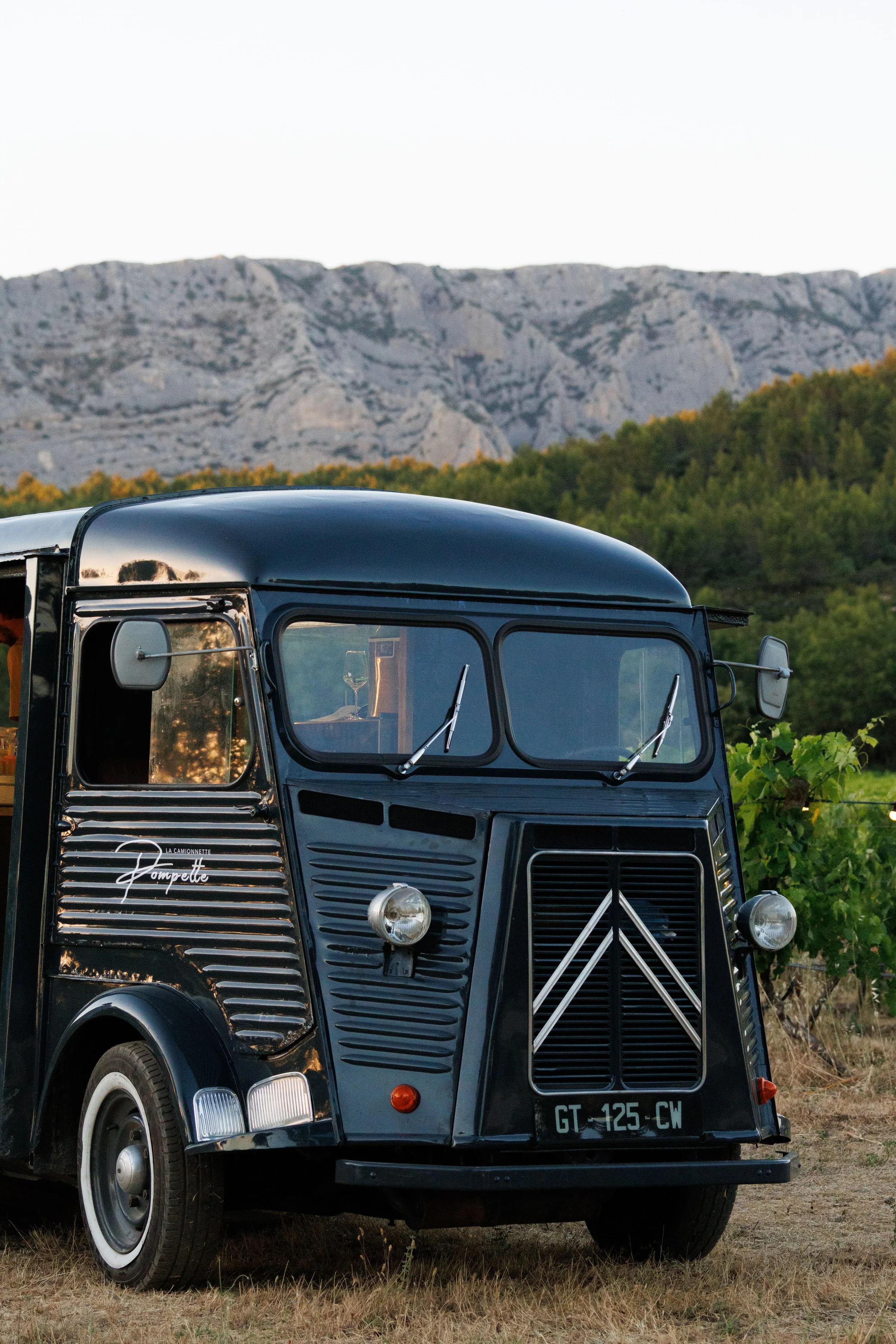 Un food truck noir avec un menu écrit à la craie, stationné dans un espace extérieur où des gens sirotent des boissons, assis sur des chaises jaunes et des tables, entourés de verdure et de jeunes arbres.