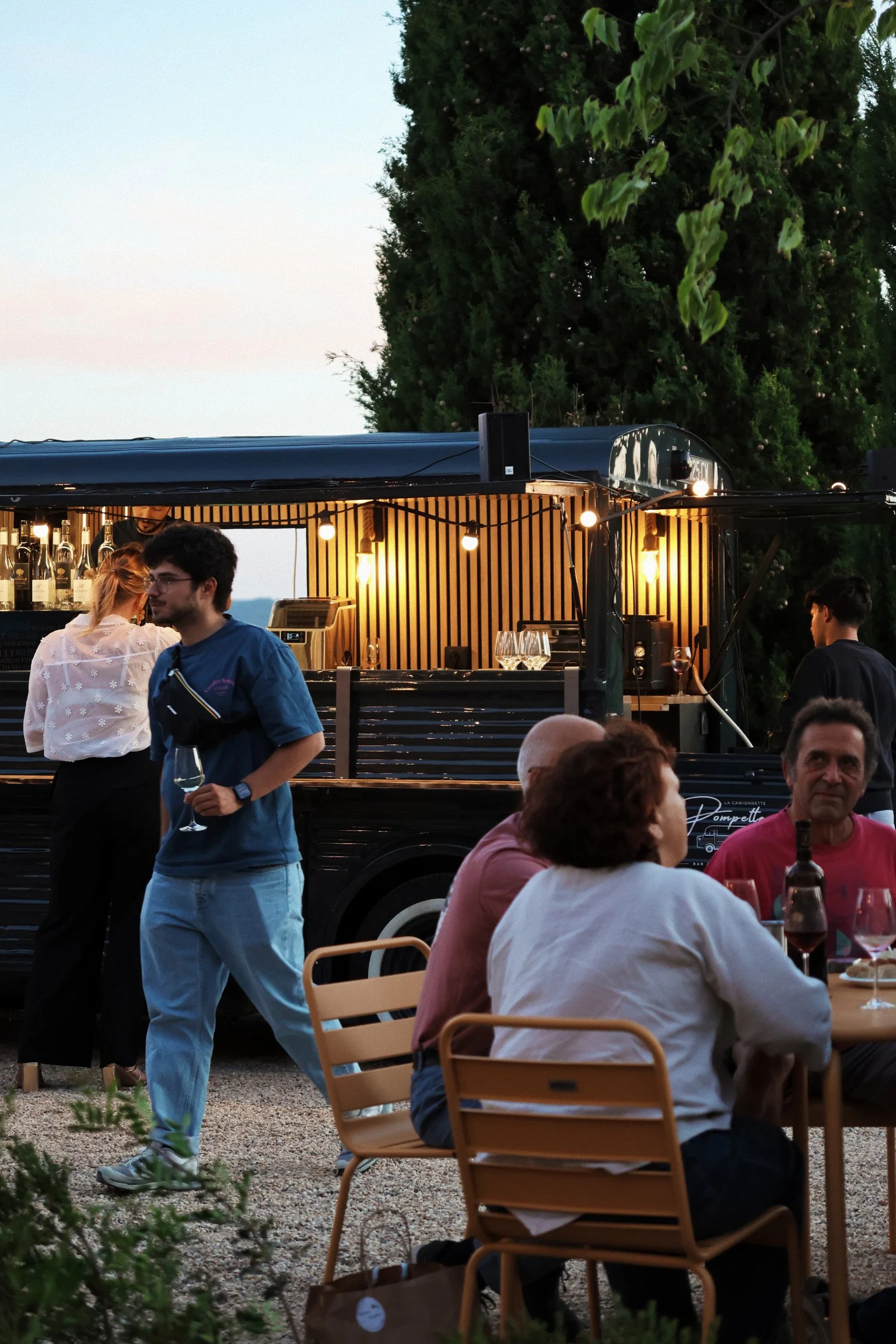 Une scène extérieure avec des personnes assises autour d'une table, un homme tenant un verre de vin, et un bar avec éclairage chaleureux en arrière-plan, entouré d'arbres.