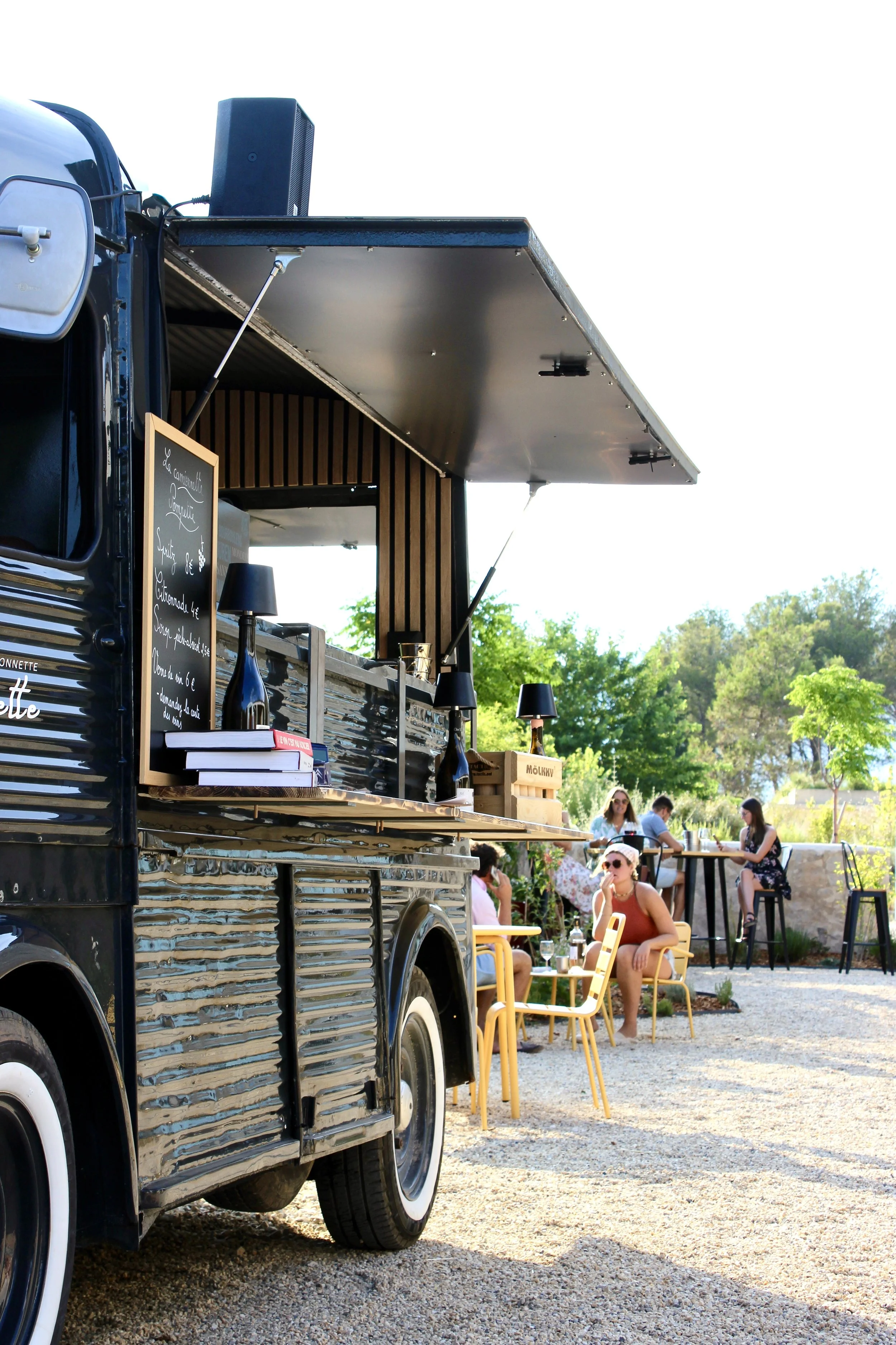 Un food truck noir avec un menu écrit à la craie, stationné dans un espace extérieur où des gens sirotent des boissons, assis sur des chaises jaunes et des tables, entourés de verdure et de jeunes arbres.