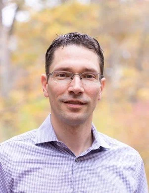 A man with short dark hair and glasses, wearing a light purple button-up shirt, standing outdoors with blurred autumn trees in the background.