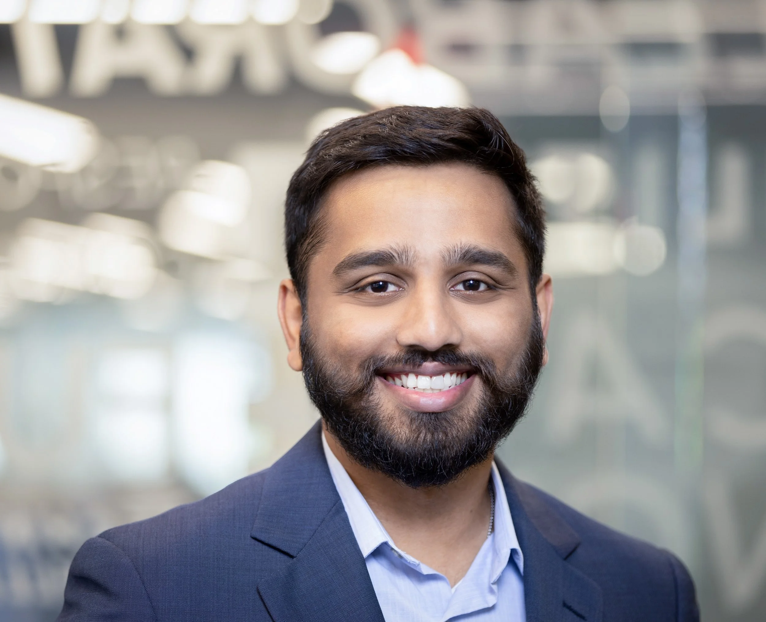 A smiling man in a blue suit and light blue shirt, with short dark hair and a beard, standing in a modern office or corporate environment with blurred background lights and glass.