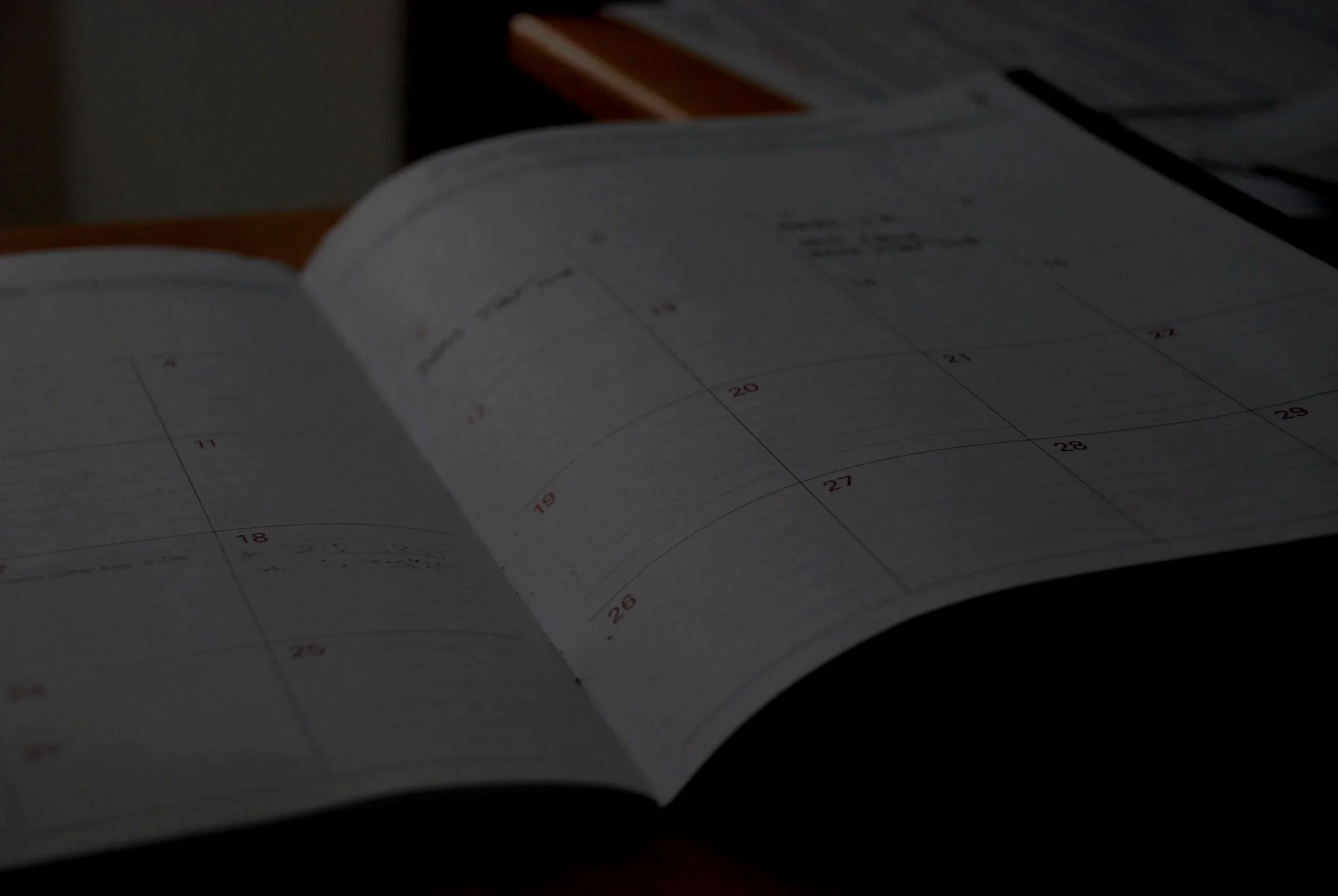 A close-up photograph of a printed calendar, with some days marked and others blank, placed on a dark surface with a small portion of a book or paper in the background.