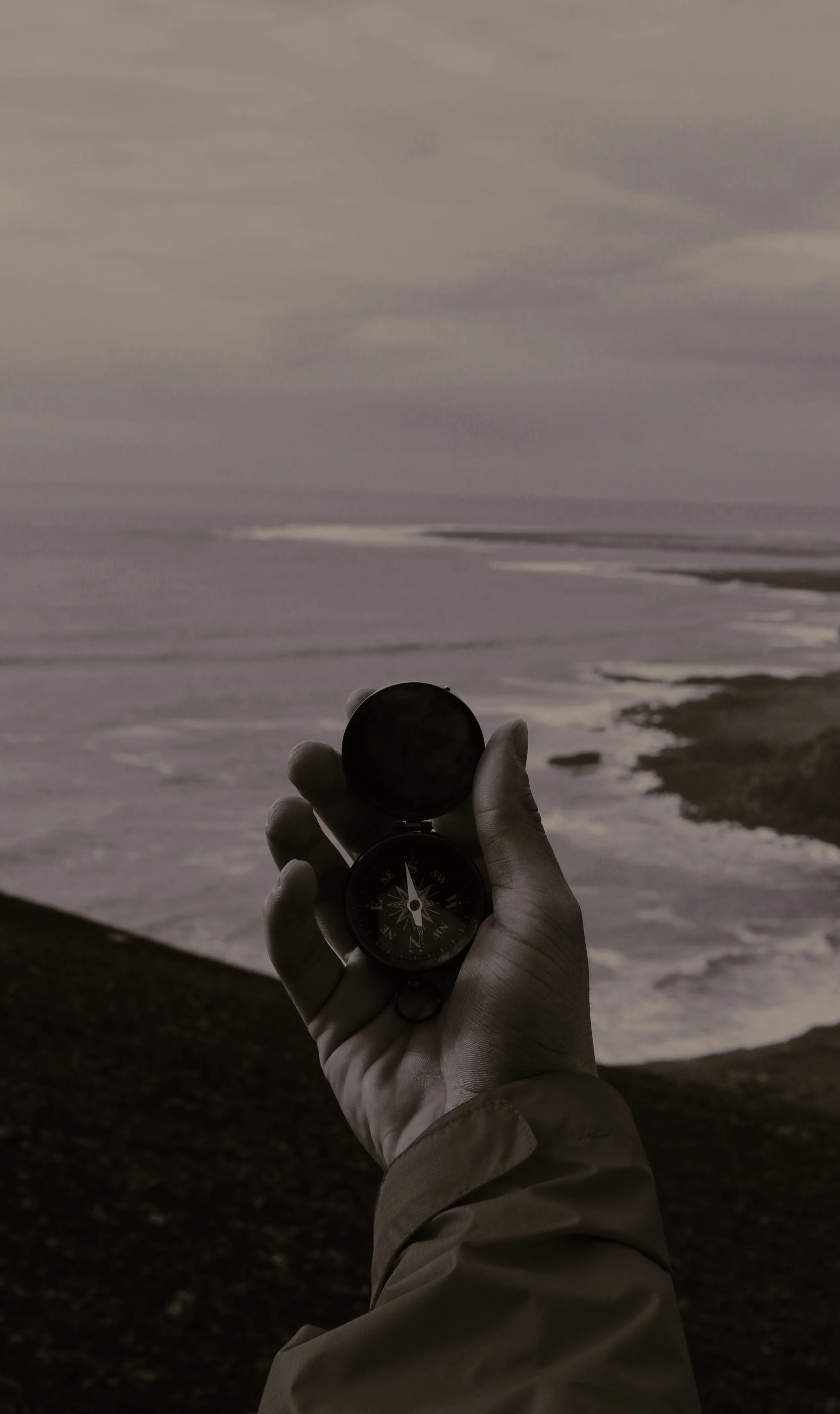 A person holding a compass with a beach and ocean in the background during overcast weather.