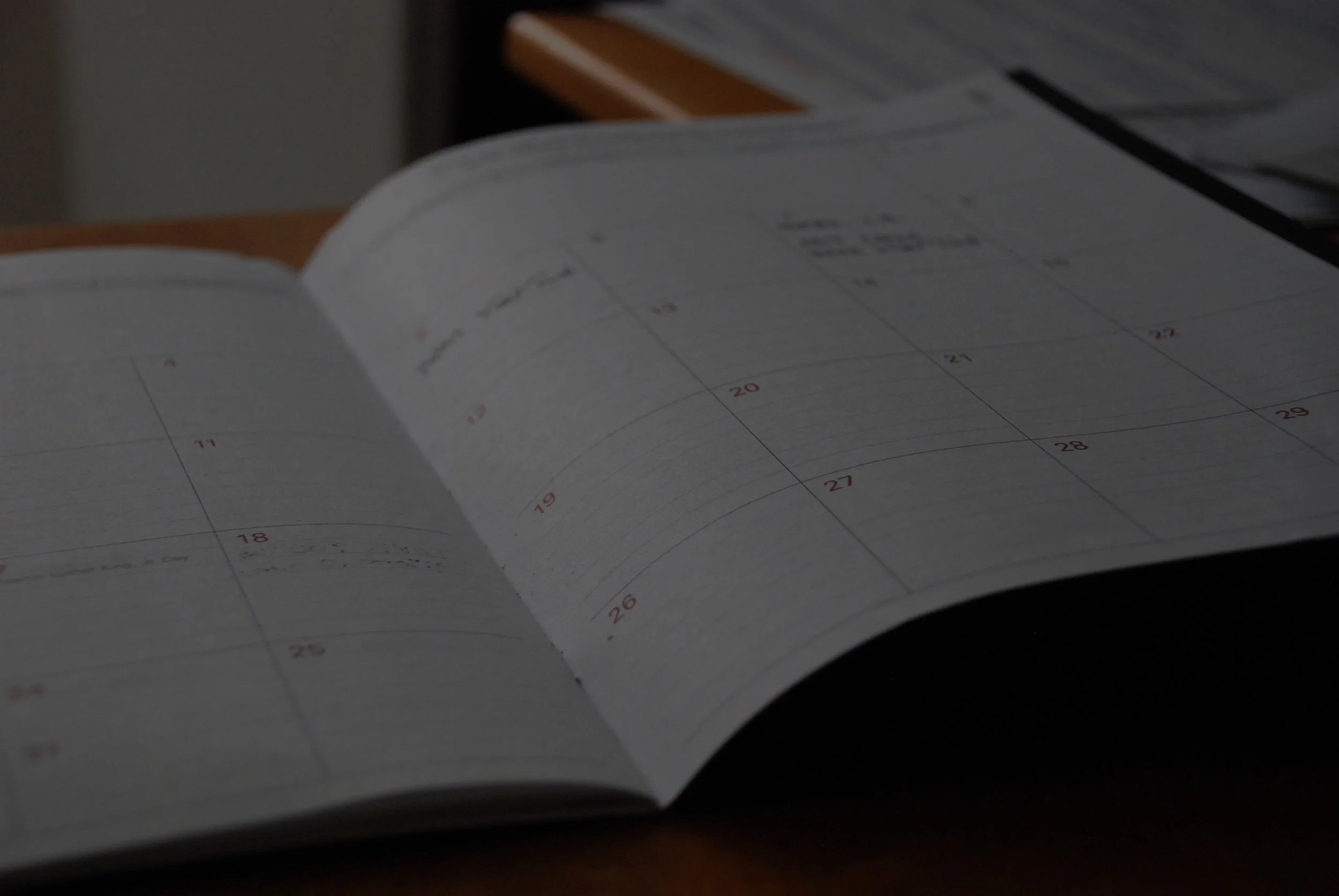 An open planner or calendar on a desk showing dates and days, with a notebook and paper elsewhere on the desk.