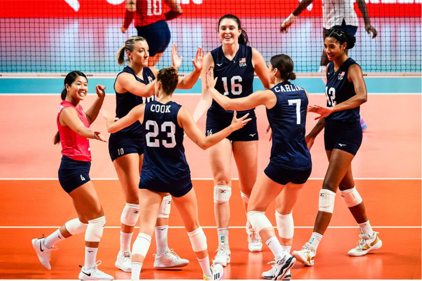 A group of female volleyball players celebrating on an indoor court, wearing navy blue and pink jerseys, with a volleyball net in the background.