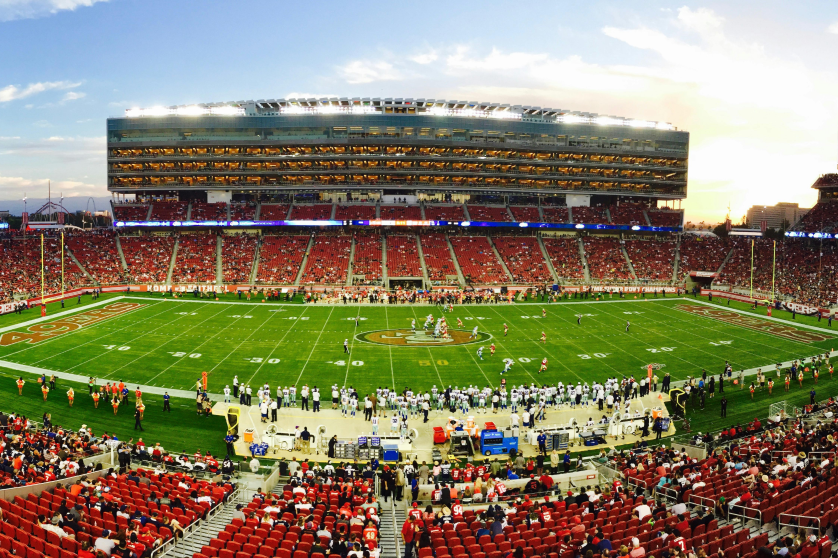 An outdoor football stadium filled with spectators, with players on the field and a sunset in the background.