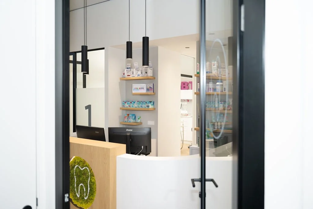 Ingang en balie van Mondzorg Voorne in Hellevoetsluis met receptie, computers, houten en glazen elementen, en mondzorgproducten op de planken.