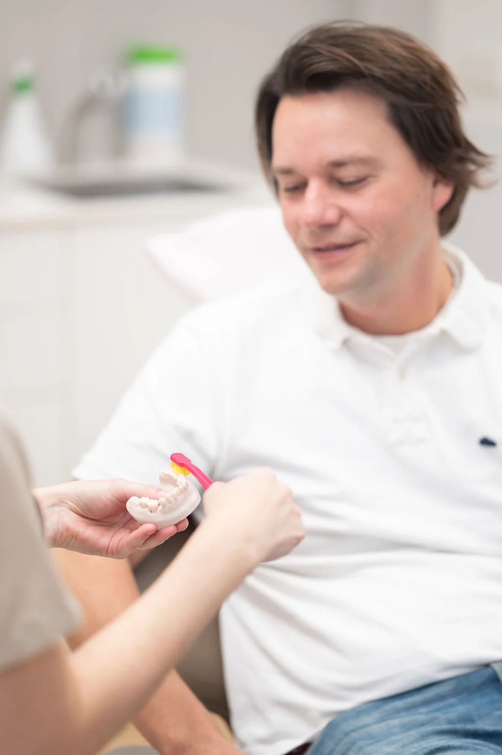 Een man zit bij mondhygienist praktijk in Hellevoetsluis, terwijl een mondhygiënist van Mondzorg Voorne in Oostvoorne een modelkaak en tandenborstel vasthoudt om mondverzorging uit te leggen.