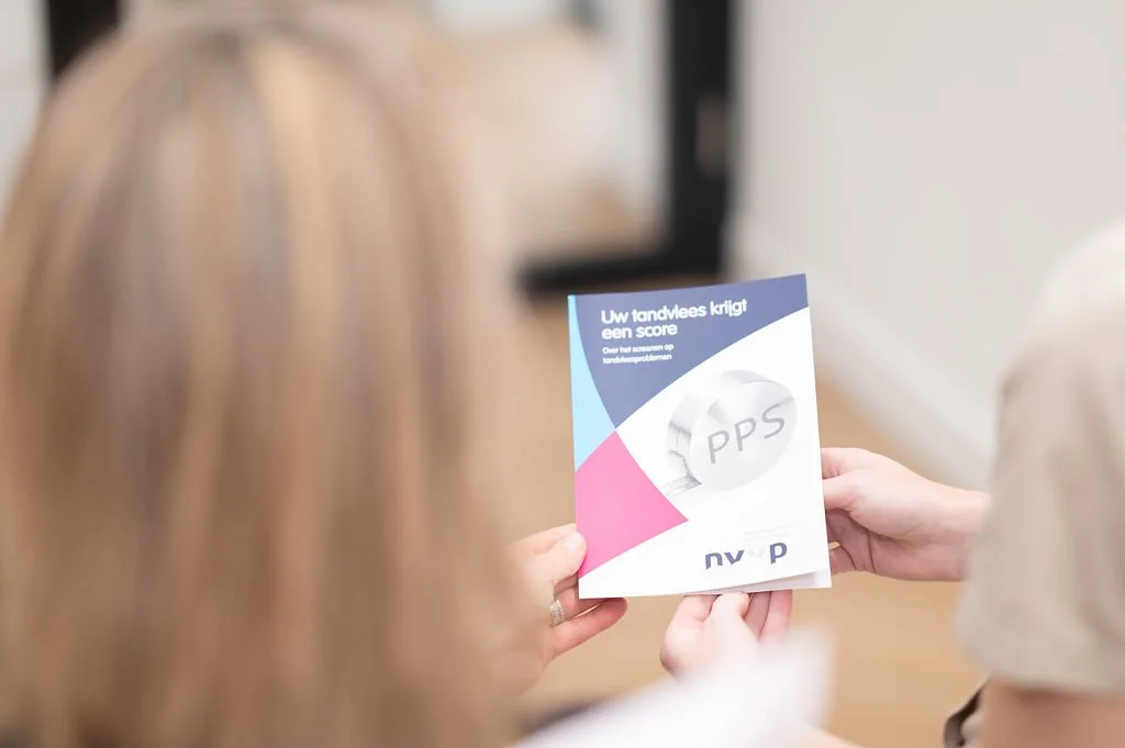Mondhygienist en cliënt bekijken een informatief blad over tandvleesproblemen bij Mondzorg Voorne in Hellevoetsluis/Oostvoorne, met de tekst “Uw tandvlees krijgt een score” en een afbeelding van een tand met “PPS” erop.