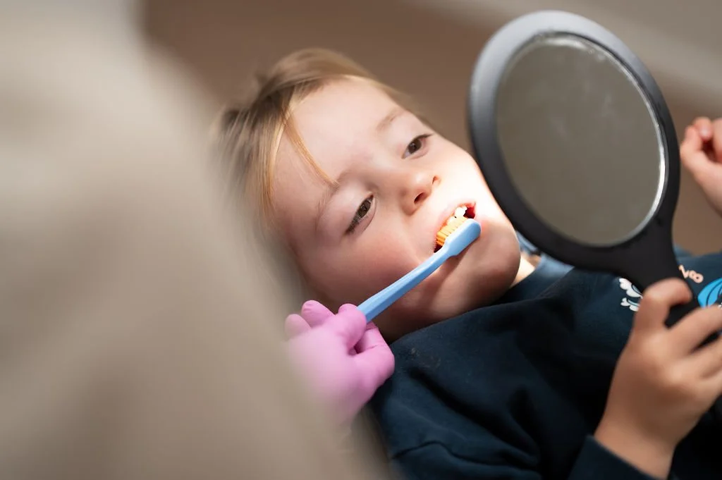 Kind zit op een stoel en kijkt in de spiegel terwijl een mondhygiënist van Mondzorg Voorne in Oostvoorne en Hellevoetsluis op de achtergrond advies geeft.
