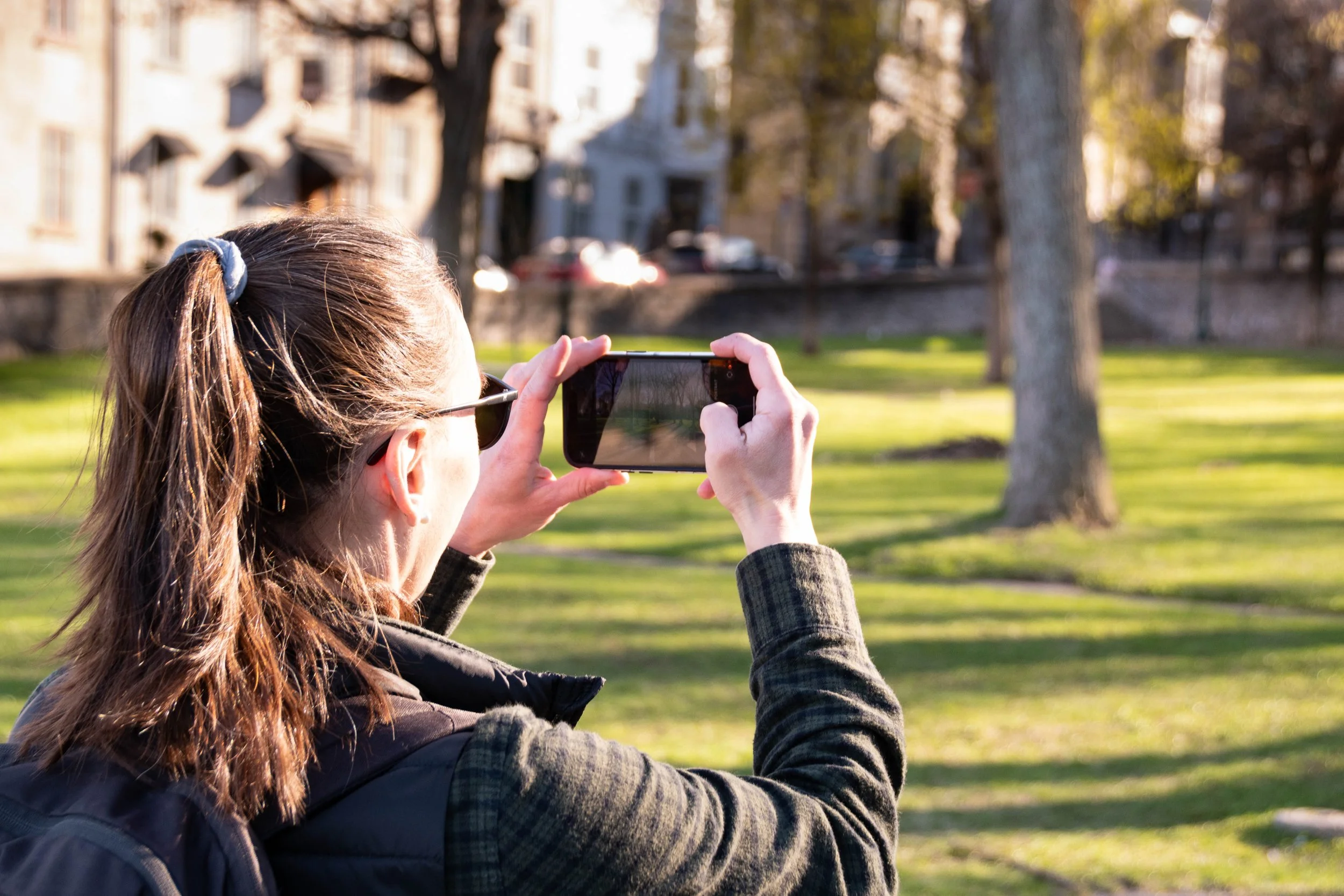 A woman with brown hair, wearing sunglasses and a backpack, takes a photo with her smartphone in a park with trees and buildings in the background.