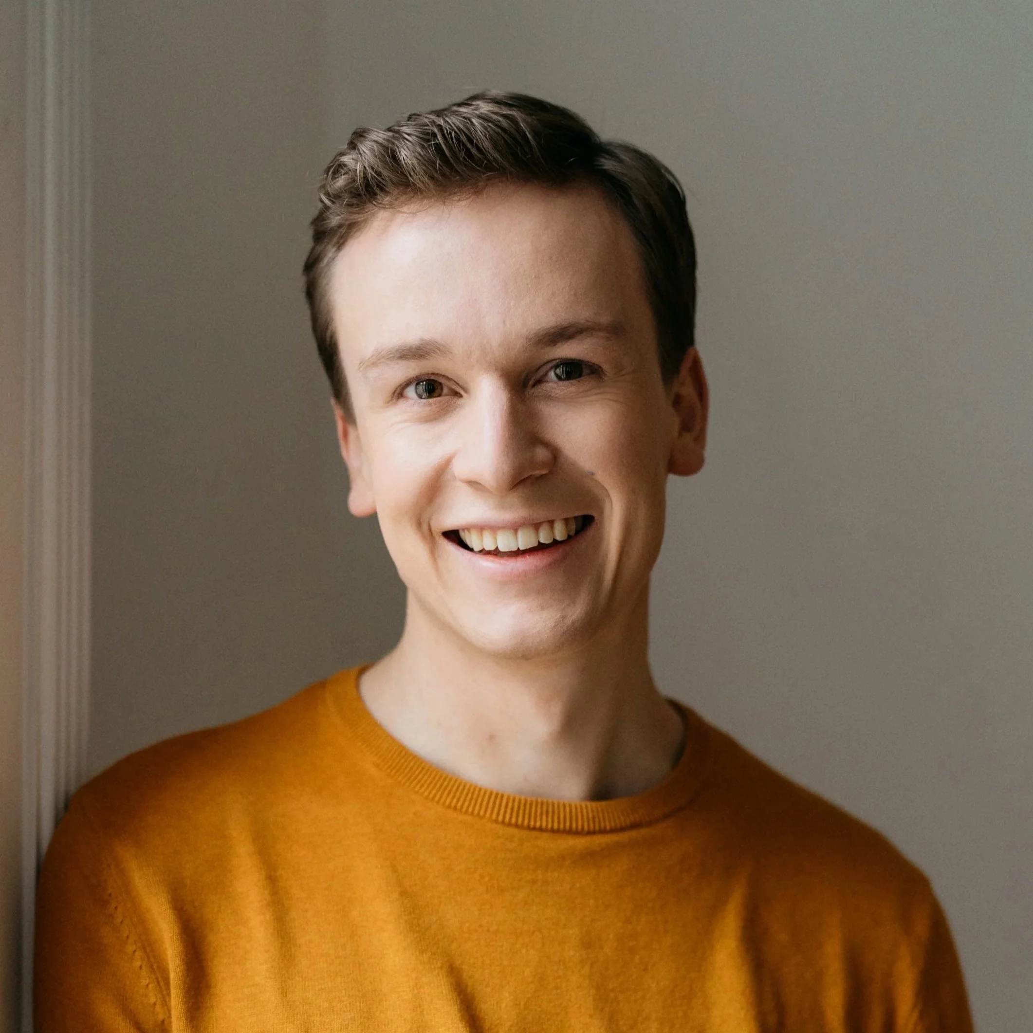 Nahaufnahme eines lachenden Mannes mit braunen Haaren, die an den Seiten kurz und oben länglich sind, trägt einen senf-farbenen Pullover.