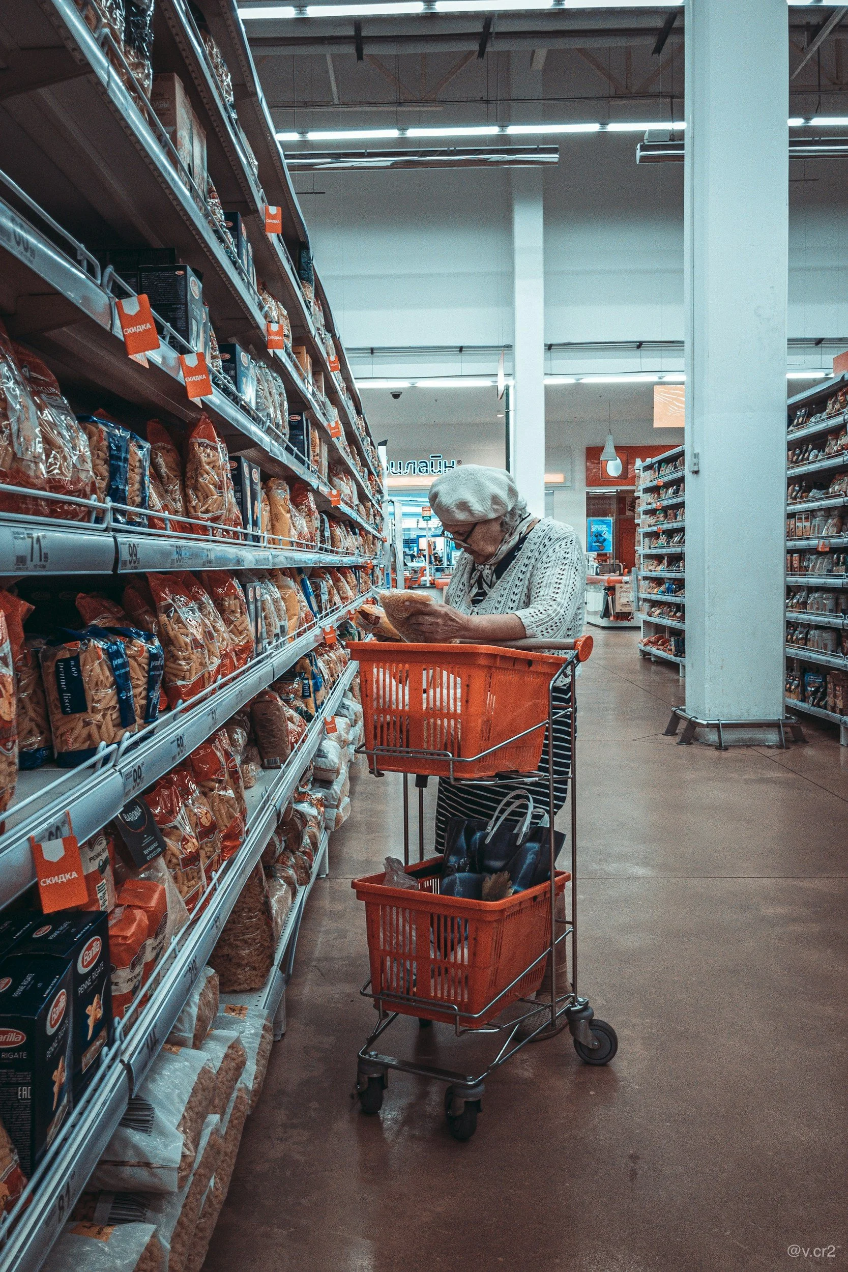 An elderly woman shopping in a grocery store aisle, examining a package of food, with an empty shopping cart beside her.