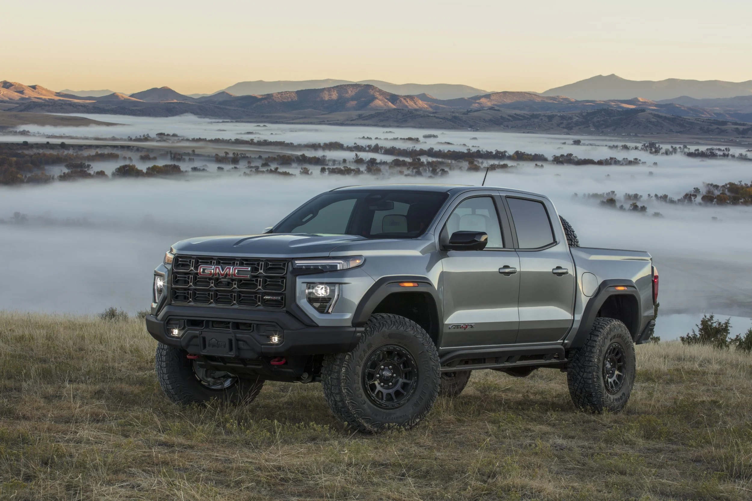Gray GMC Sierra pickup truck parked on a grassy hill with a foggy landscape and mountains in the background.