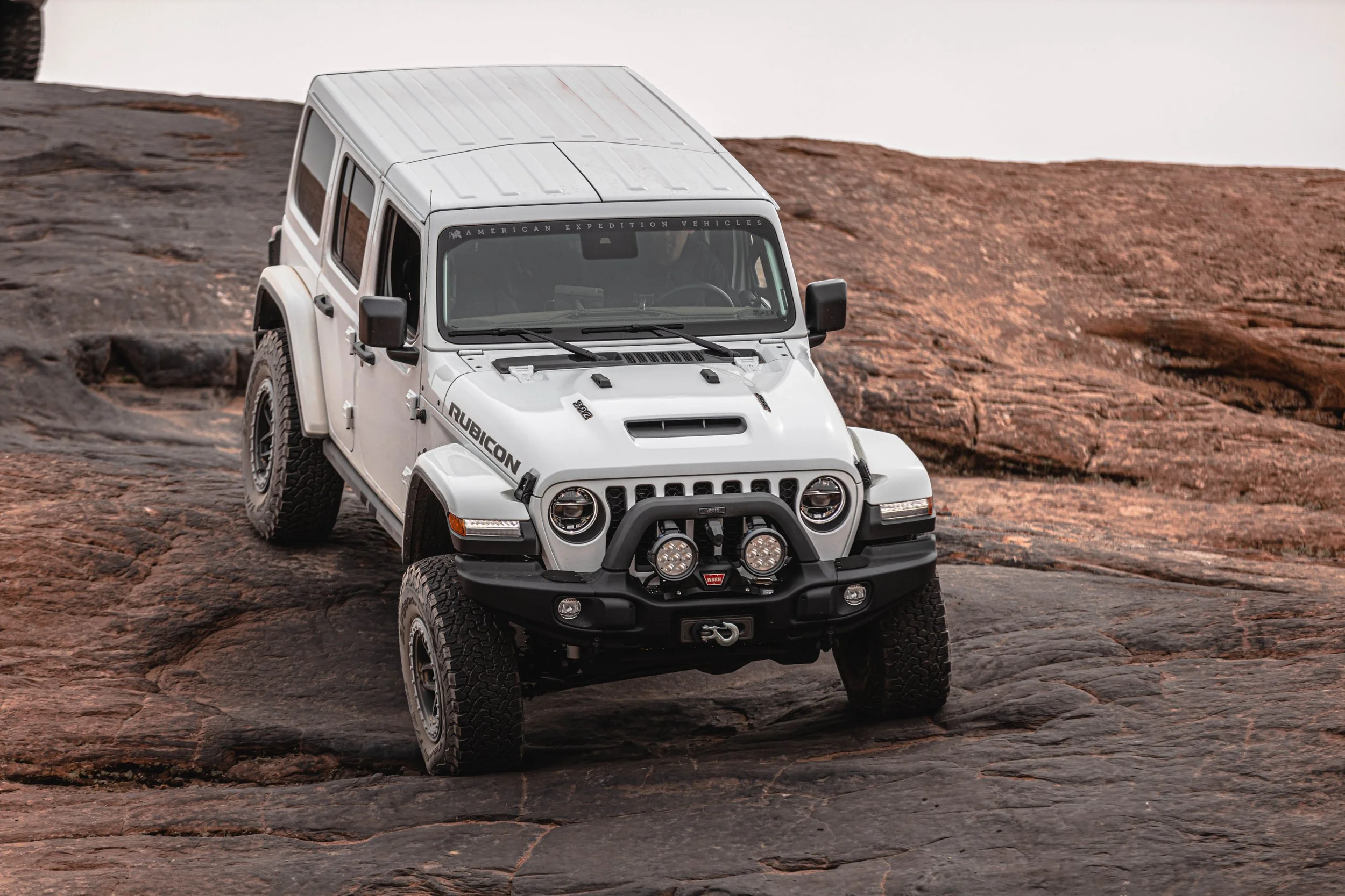 A white off-road toy Jeep Rubicon with black accents climbing reddish-brown rocky terrain outdoors.