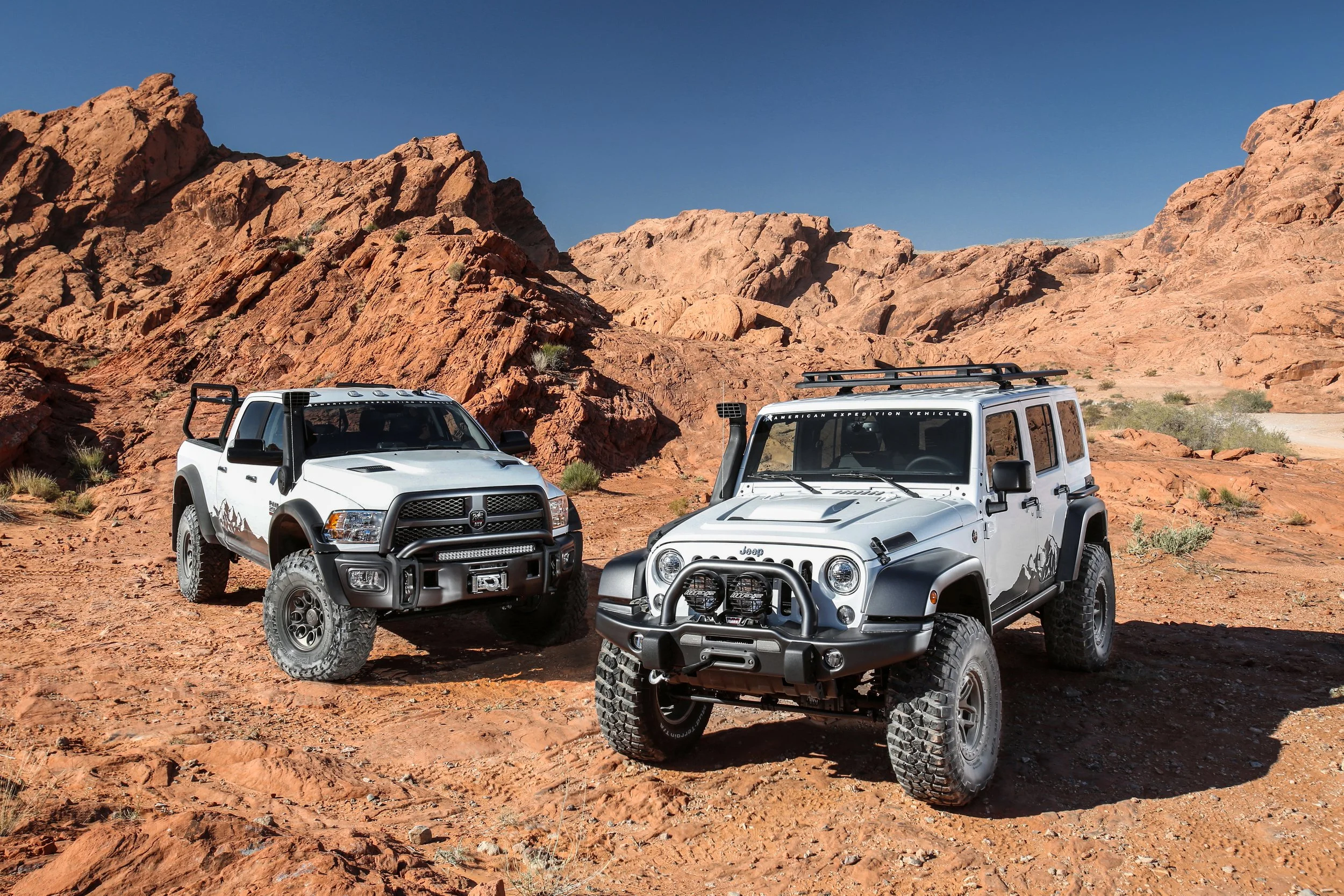 Two off-road vehicles parked on a rocky desert landscape with red rock formations and sparse vegetation under a clear blue sky.