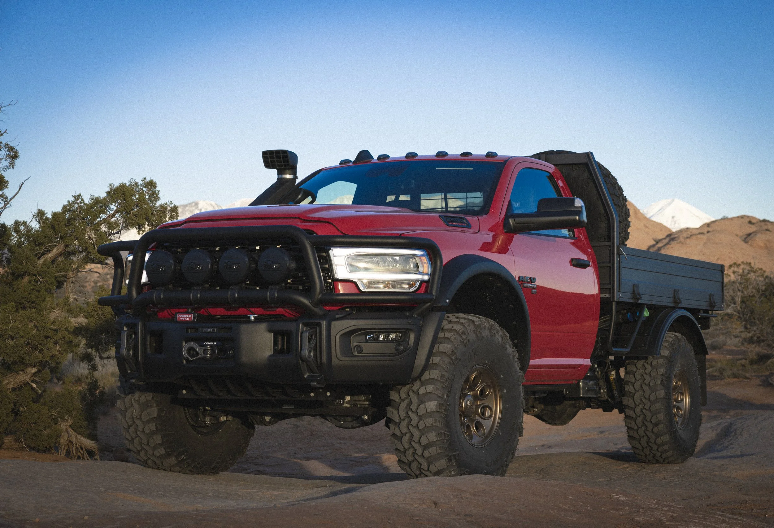 Red off-road pickup truck with large tires, black front bumper with lights, and a black cargo bed, driving on rugged terrain with mountainous background.