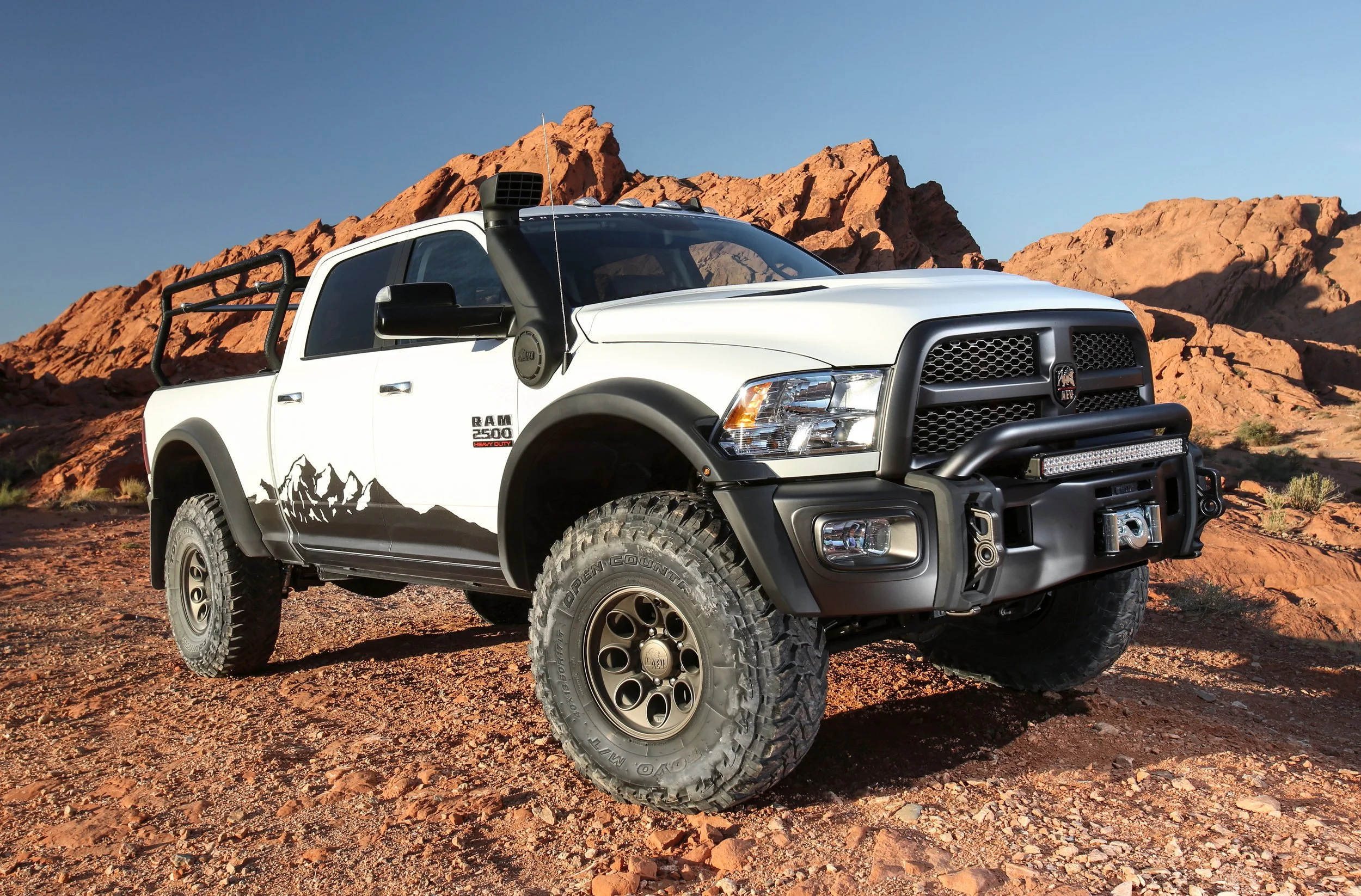 A white Ram 2500 off-road truck with mountain graphics on the side, parked on a rocky desert landscape with red rock formations in the background and clear blue sky.