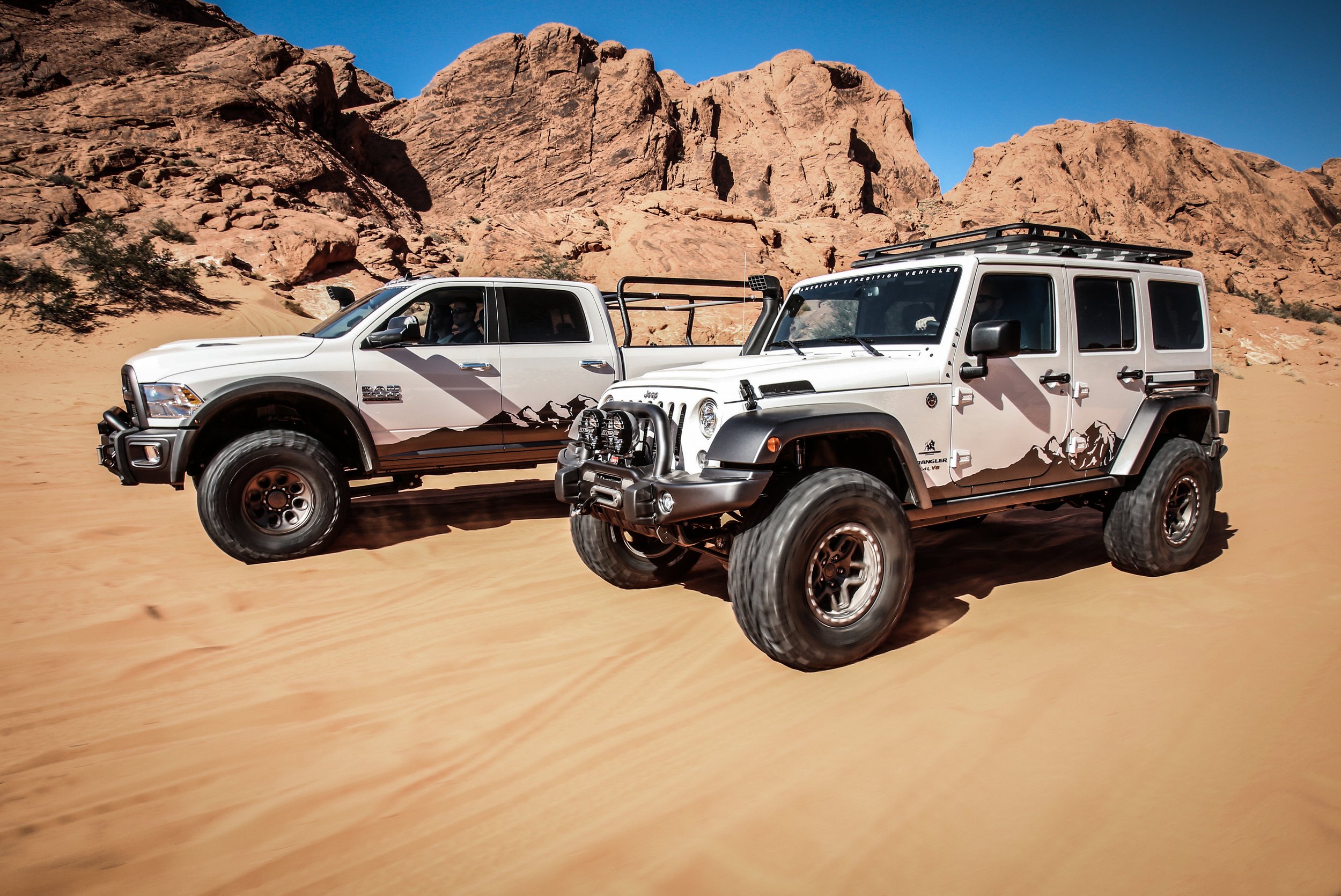 Two white off-road vehicles with mountain and desert background, featuring mountain graphics on their sides, engaged in off-road driving in a desert landscape.