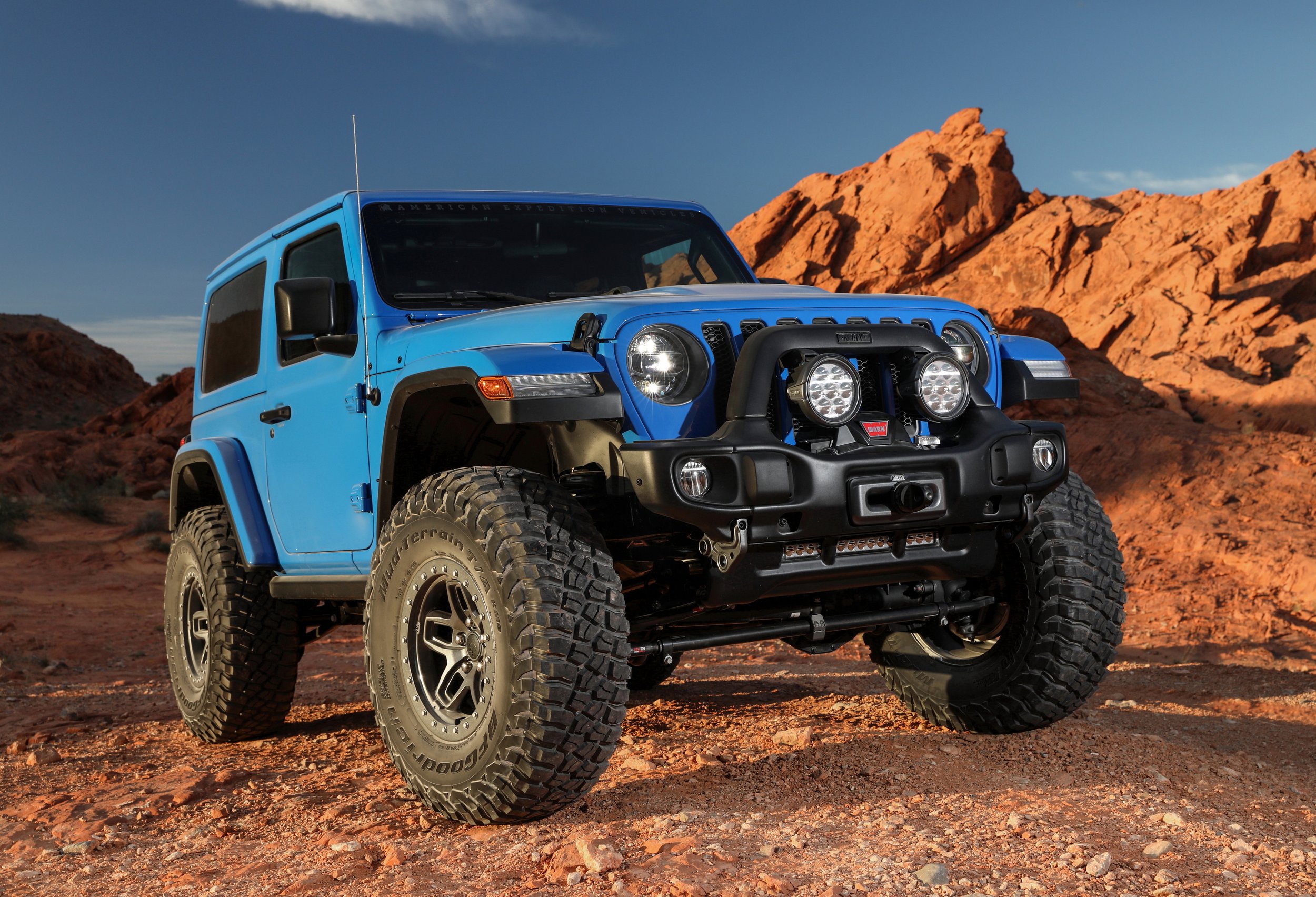 A blue off-road Jeep vehicle with large tires and additional front lights, parked on a rocky desert landscape with red rock formations in the background during daytime.