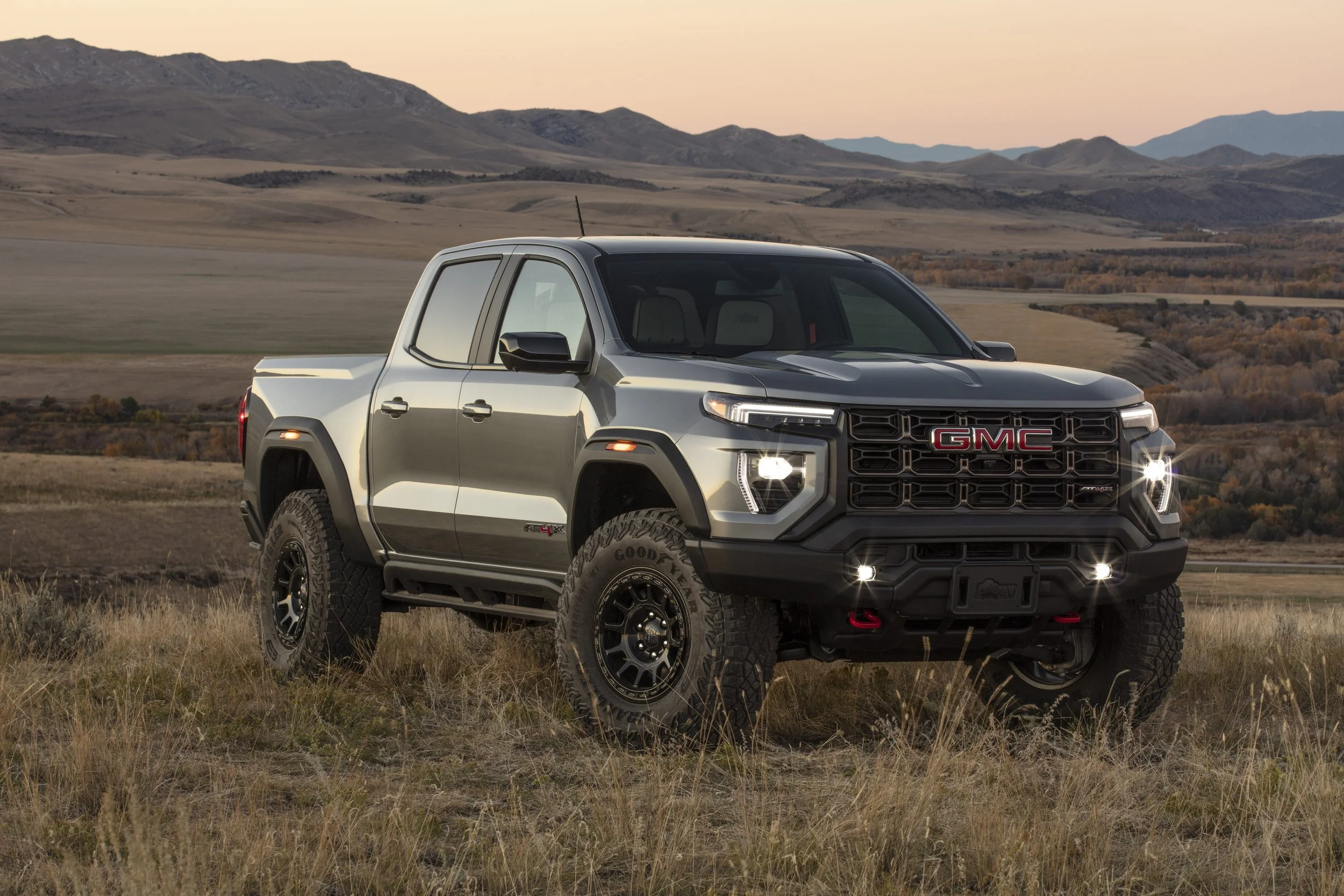 A black and silver GMC Sierra pickup truck with off-road tires parked on a grassy plain with mountains in the background at sunset.