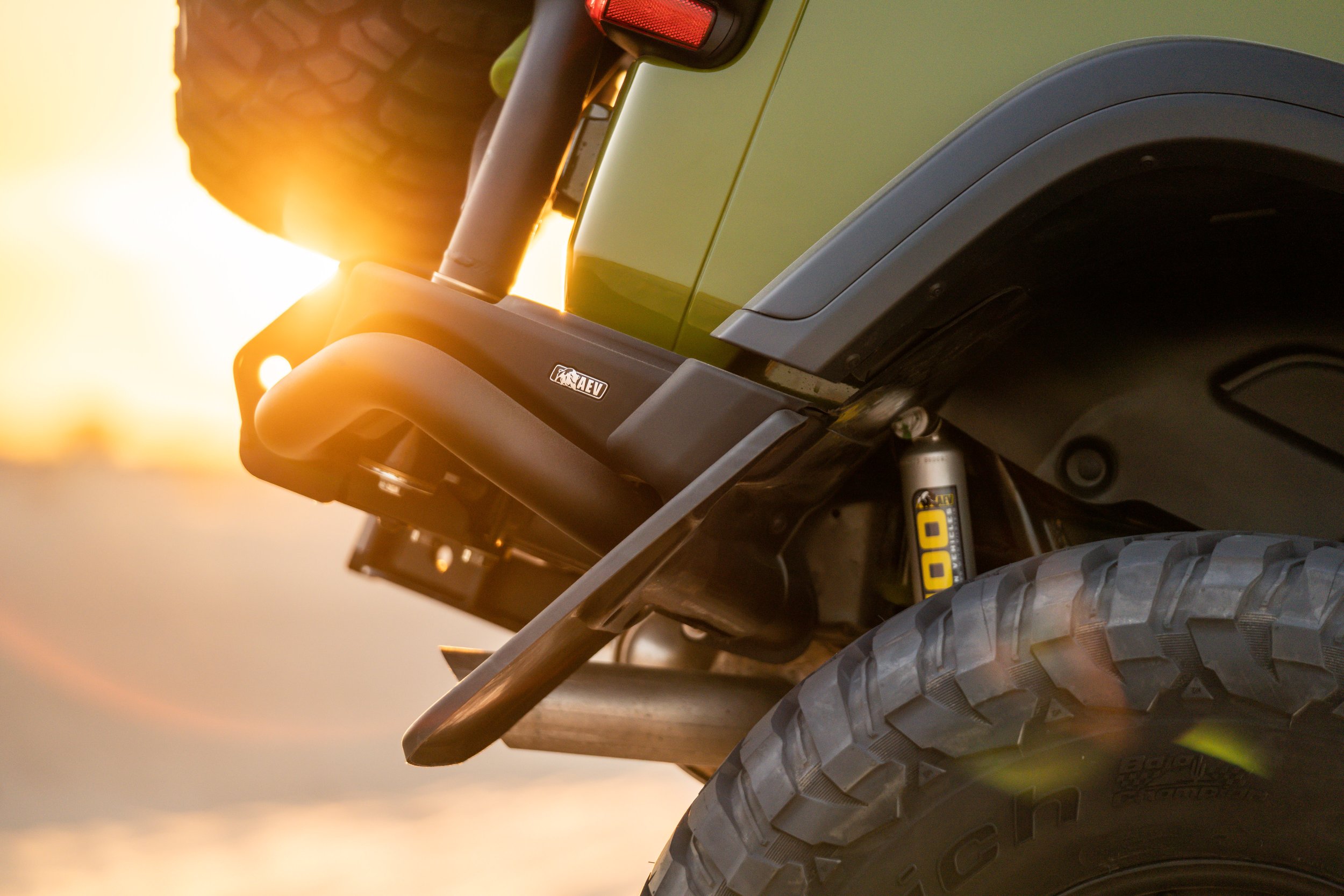 Close-up of an off-road vehicle's front tire and suspension system during sunset, highlighting rugged tires, a skid plate, and suspension components.