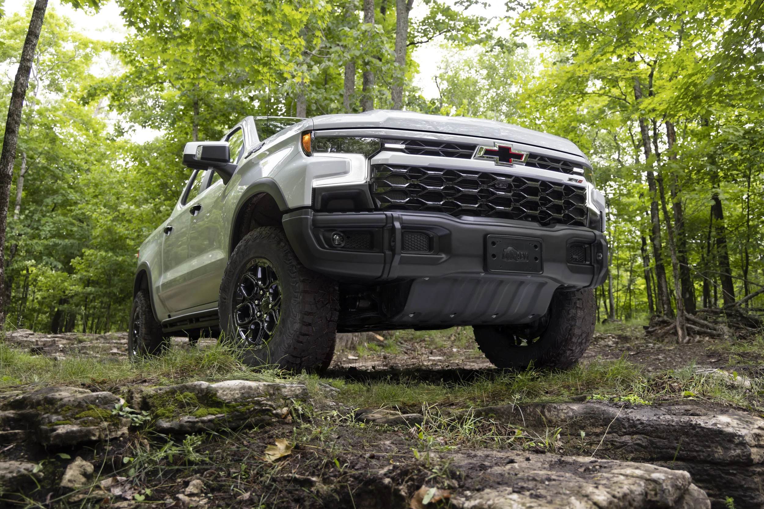 A silver Chevrolet pickup truck with off-road tires parked on a rocky trail in a dense green forest.