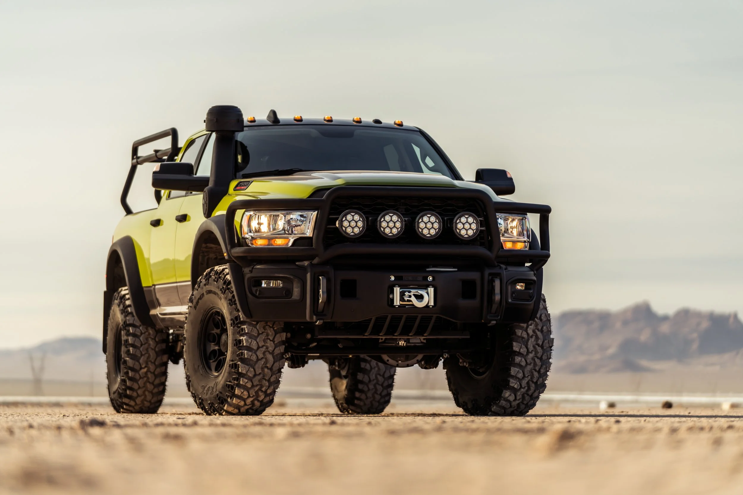 A yellow and black off-road pickup truck with large tires and a front bumper guard, parked on a desert landscape with mountains in the background.