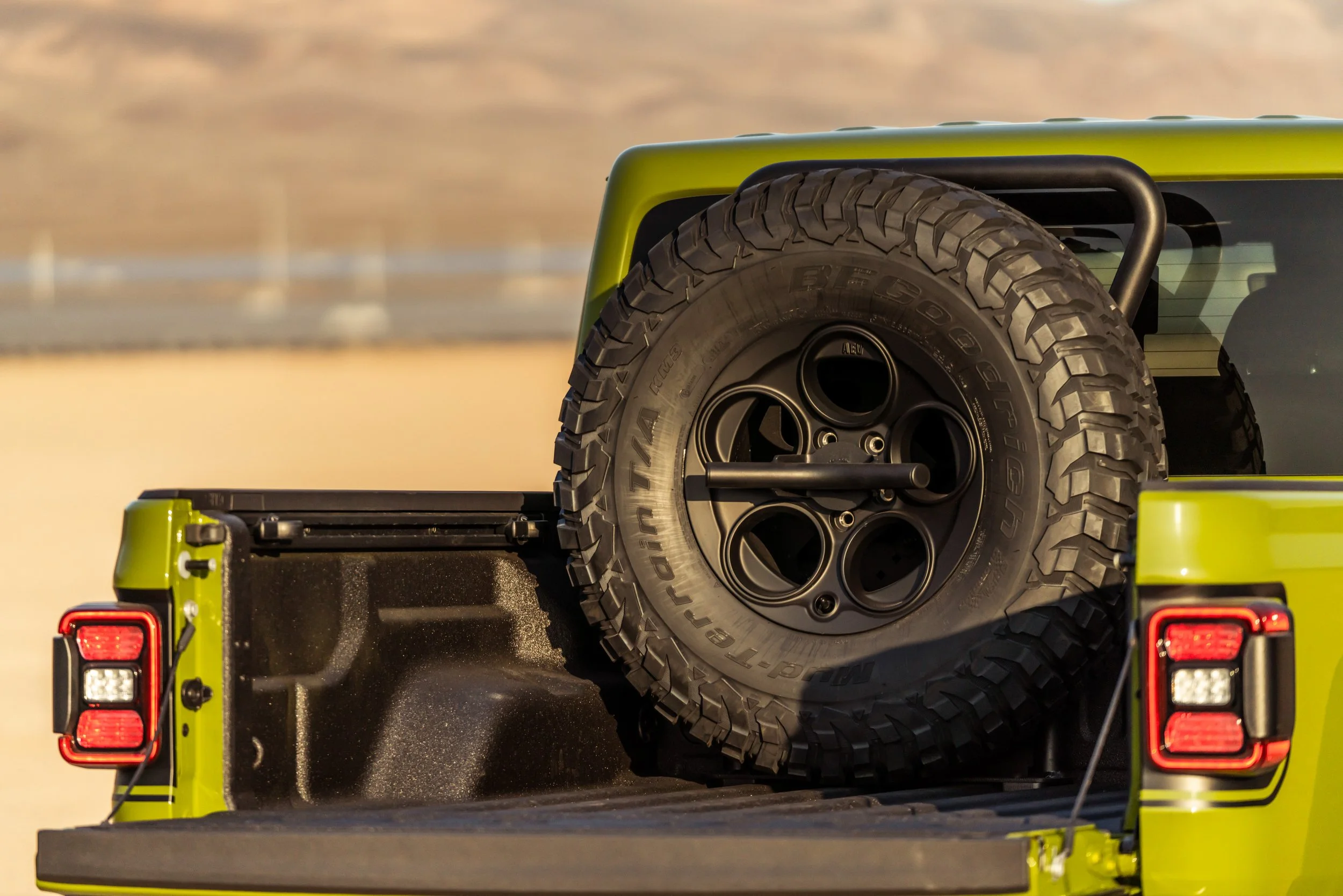 A spare tire mounted in the bed of a lime green pickup truck with black taillights, set against a desert landscape background.