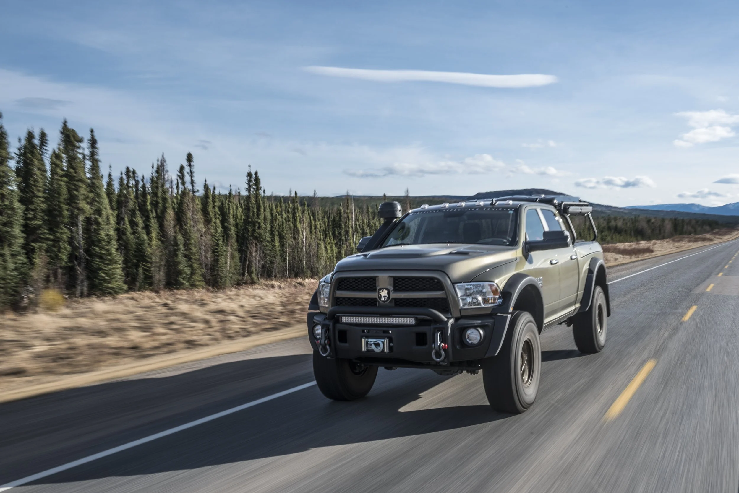 A grey Dodge Ram pickup truck driving on a two-lane highway with a forested landscape and mountains in the background under a blue sky with scattered clouds.