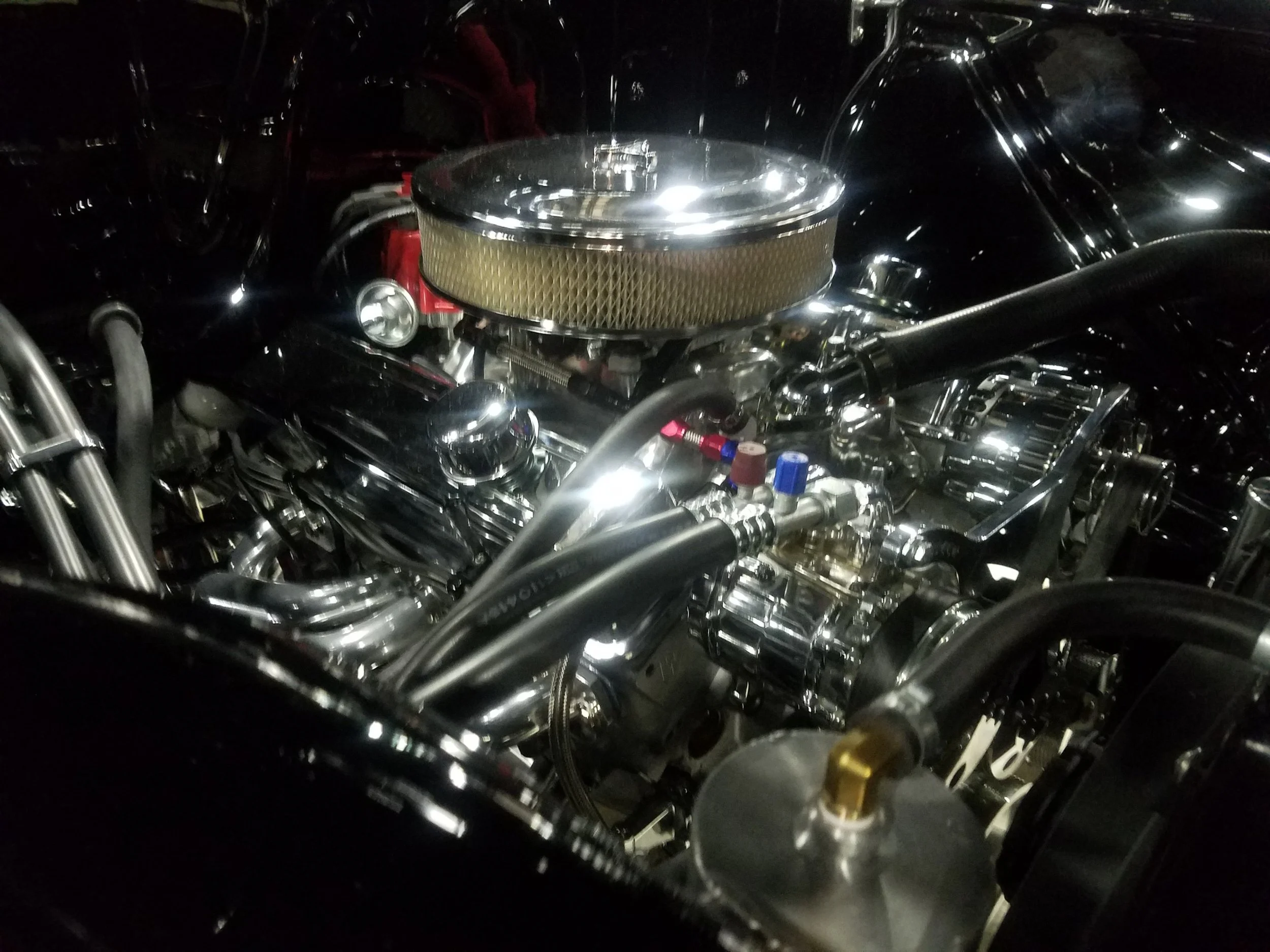 Close-up of a car engine with chrome and metallic parts, including a round air filter on top and various hoses and components.