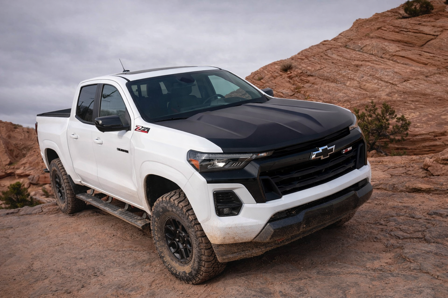 White Chevrolet pickup truck with black hood and trim, off-road tires, parked on dirt in a rocky desert landscape with red rock formations and sparse vegetation.